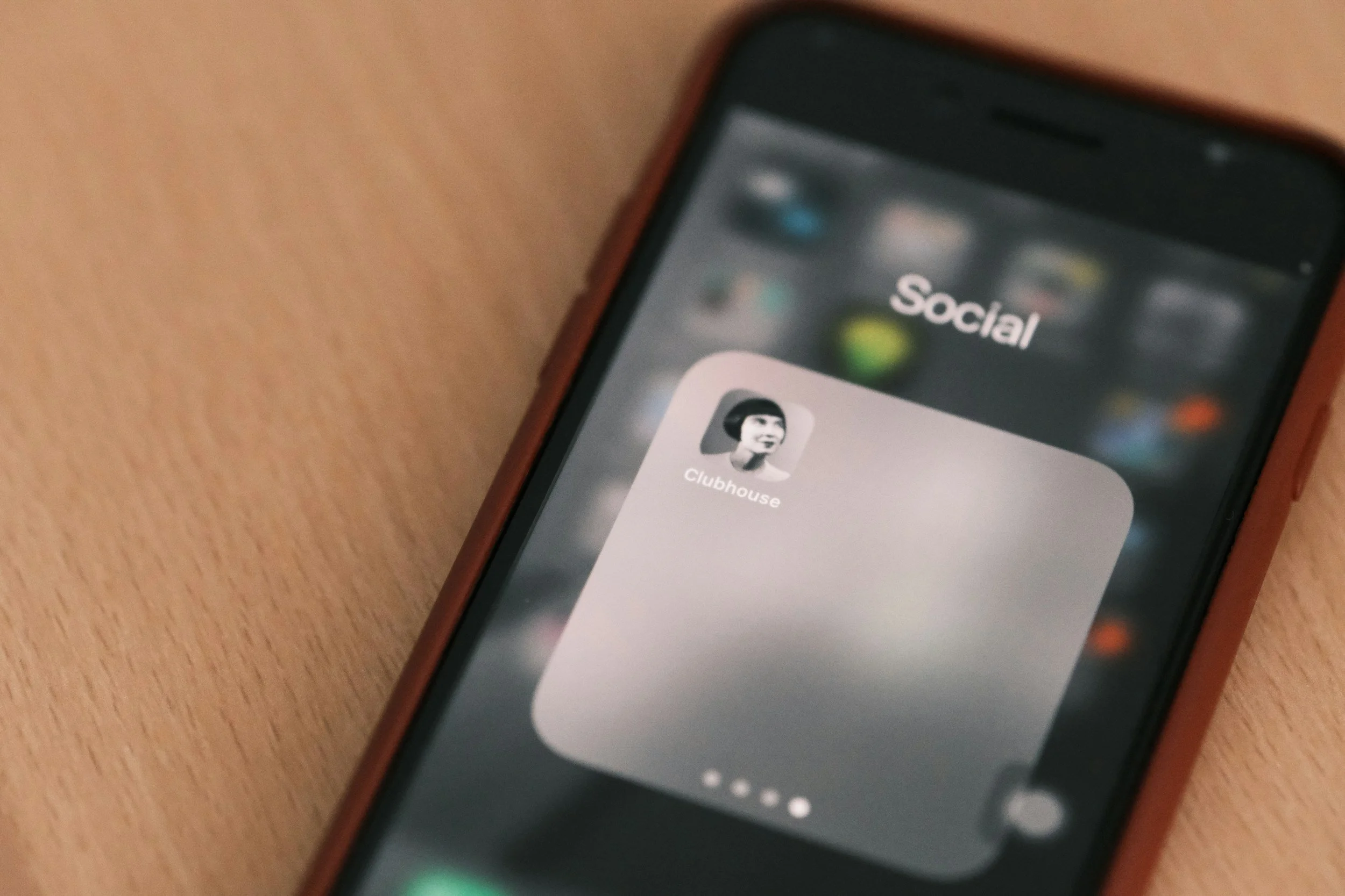 スマートフォンの画面にソーシャルフォルダ内のアプリアイコンとフォルダ名が表示されている。アイコンは白黒の女性のプロフィール写真のクラブハウスアプリ。背景は木製のテーブル。