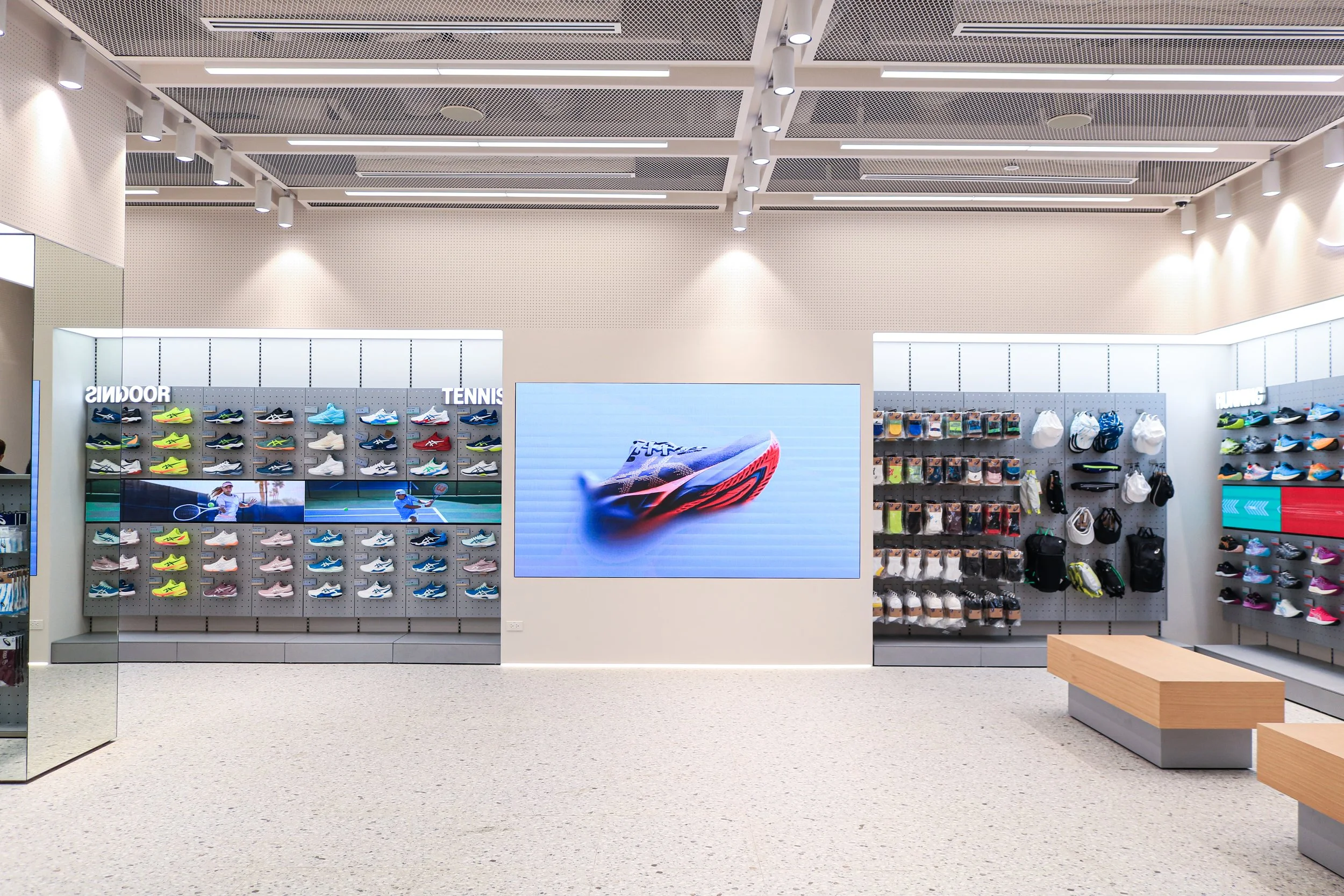 室内のスポーツ用品店で、左側にテニスシューズ、右側にバックパックや小物類が展示されている。中央にデジタルスクリーンでラッパーの新しいシューズが映し出されている。