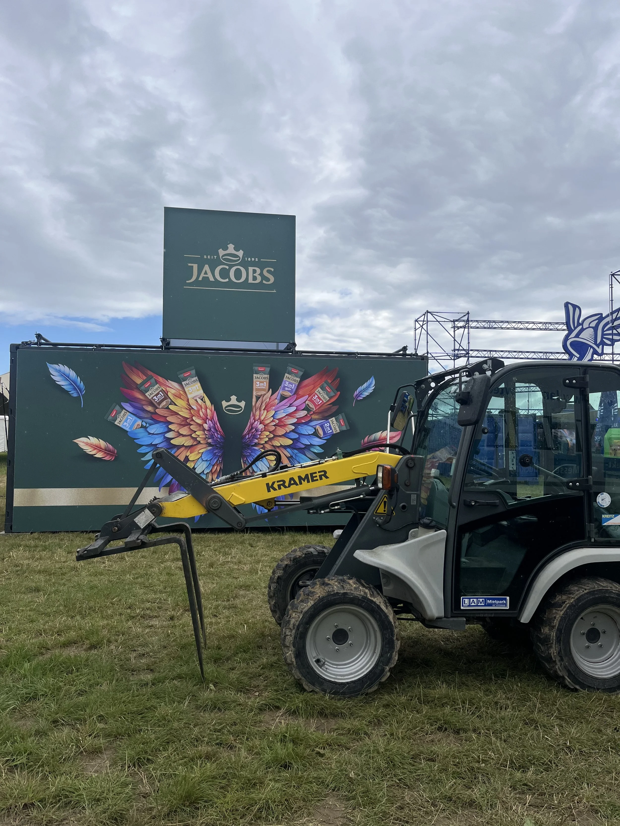 A Kramer front loader tractor parked on grass in front of a Jacob's branded billboard with a colorful angel wing design featuring various product images, under a cloudy sky.