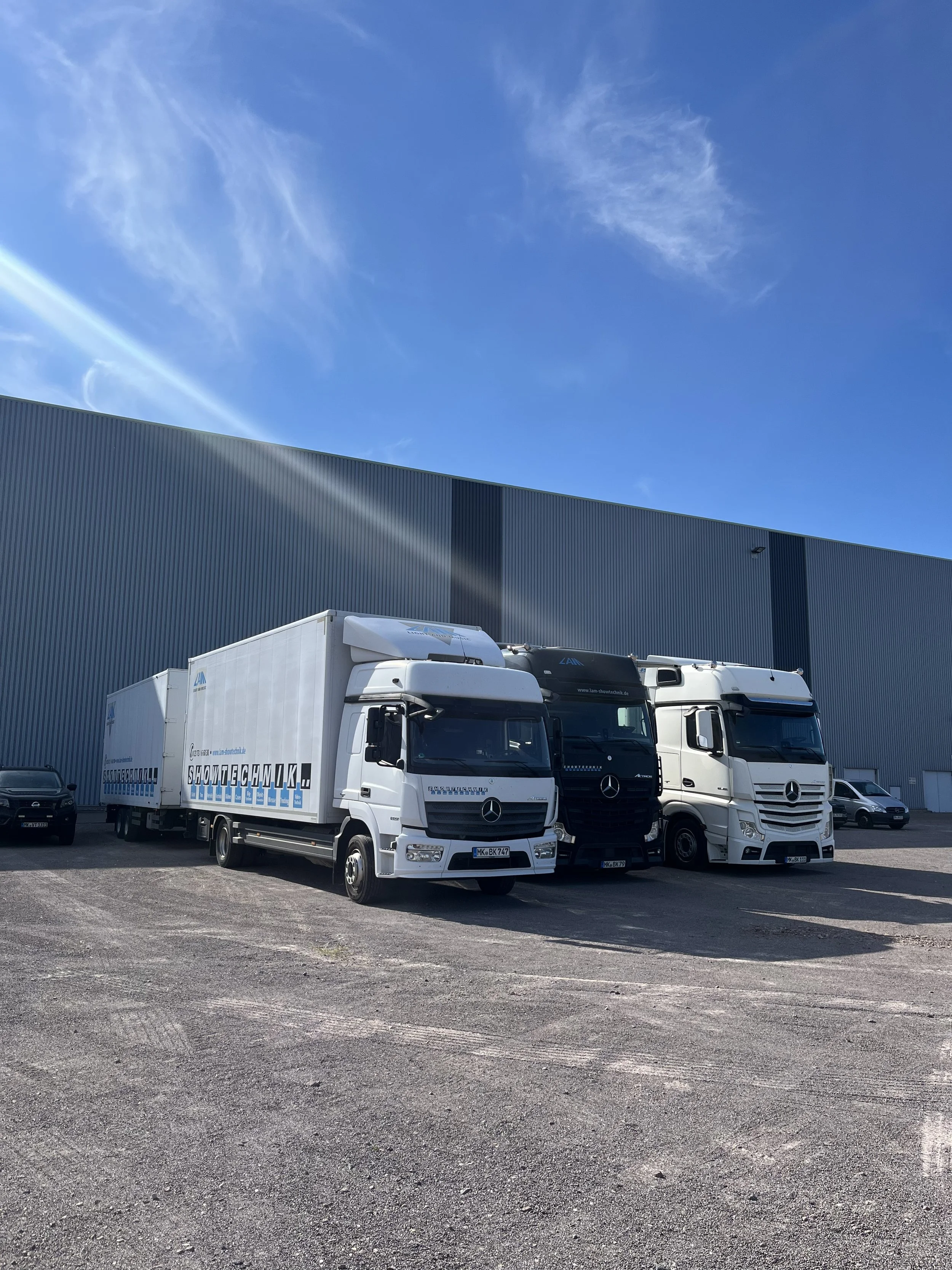 Three Mercedes-Benz trucks parked in front of a large industrial building with a blue sky overhead.