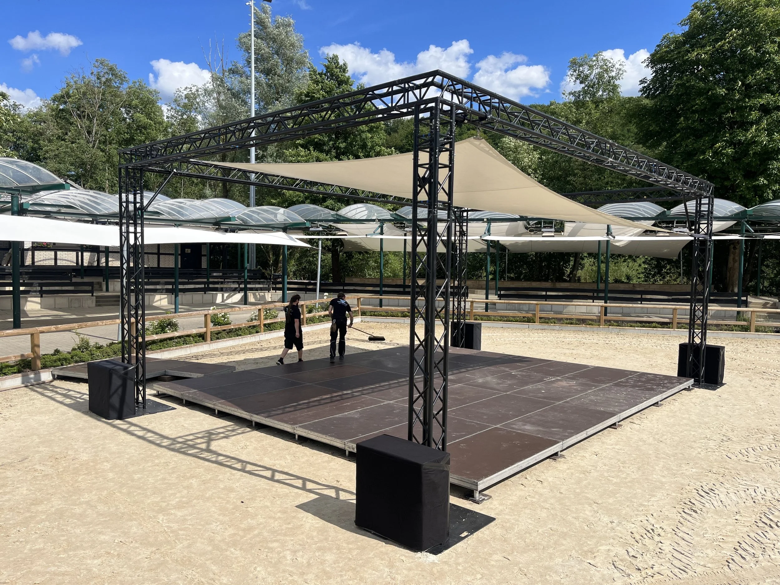 An outdoor stage setup on a dirt surface with a black metal truss frame and beige sunshade. Two people are on the stage, preparing it, with a background of trees, green foliage, and a clear blue sky with clouds.