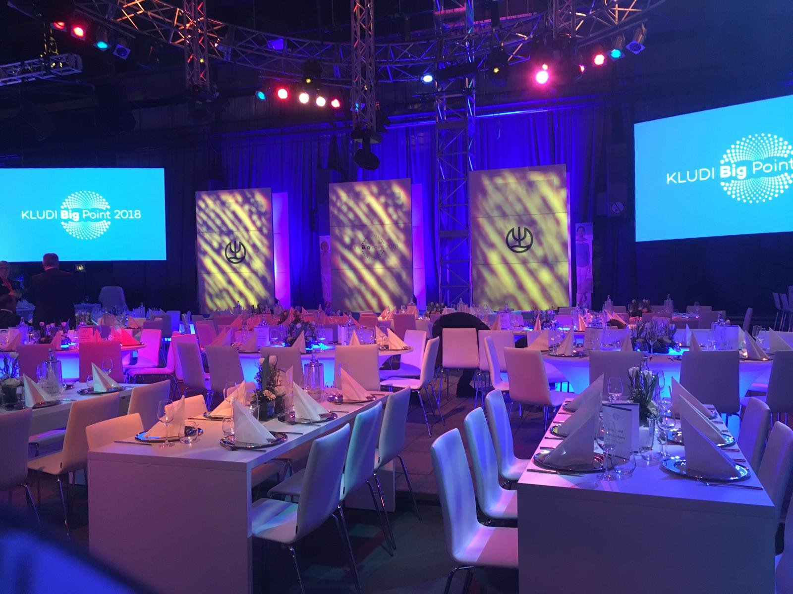 Event space set for a formal dinner at a conference with round tables, white chairs, and elegant table settings. Large screens display 'KLUDI Big Point 2018' branding. Stage with lighting and decorative panels is in the background, illuminated with p