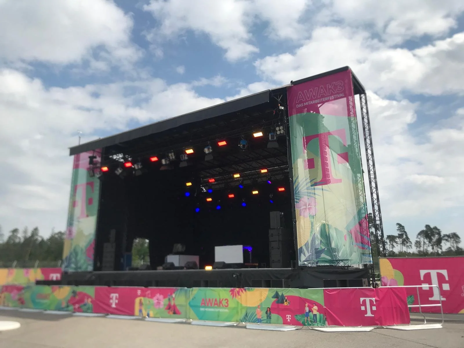 Empty outdoor stage setup for a music festival with colorful banners and a cloudy sky in the background.