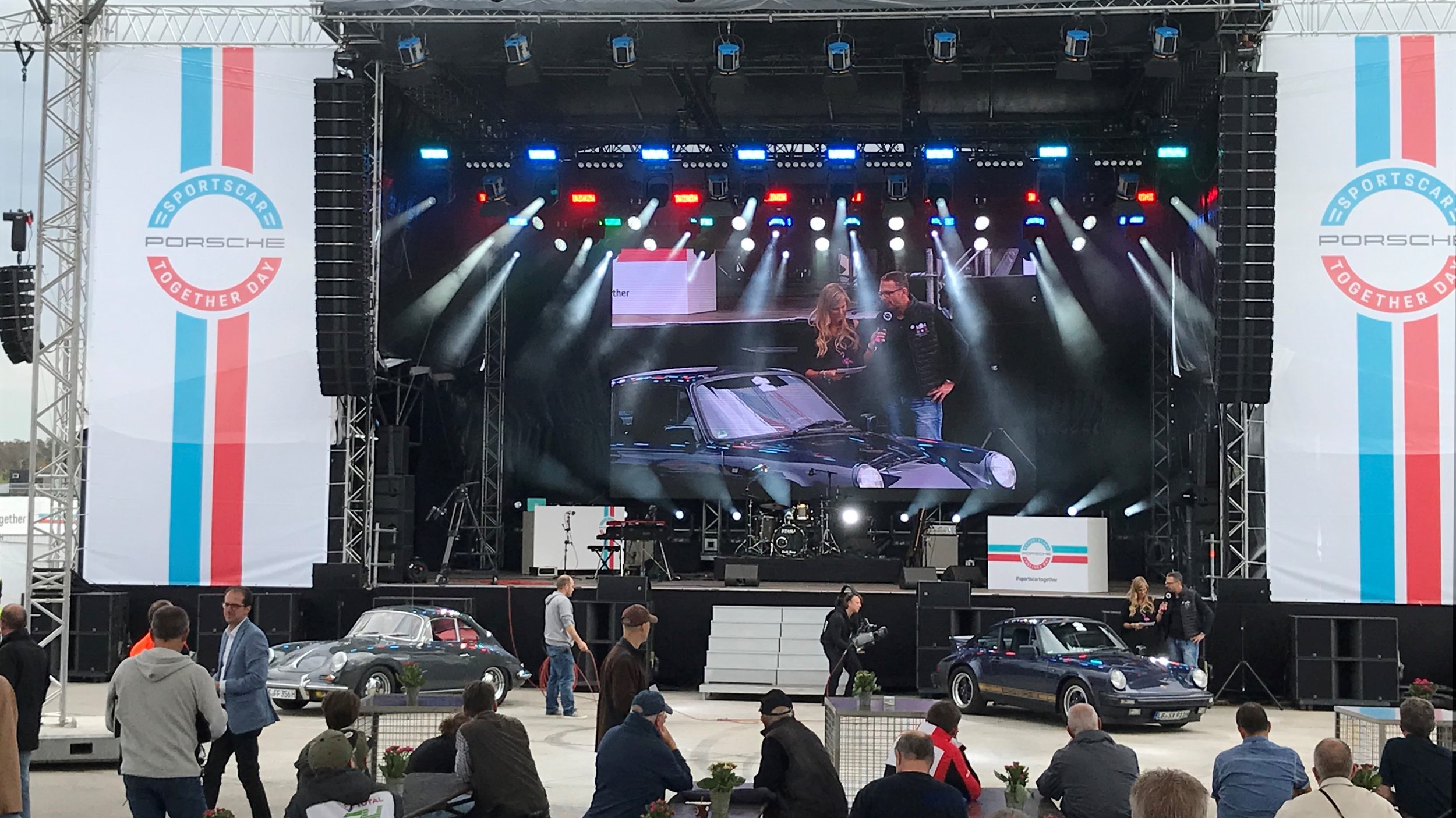 An indoor event showcasing Porsche cars, with a large stage displaying a presentation, two Porsche cars on display in front, and people gathered around.