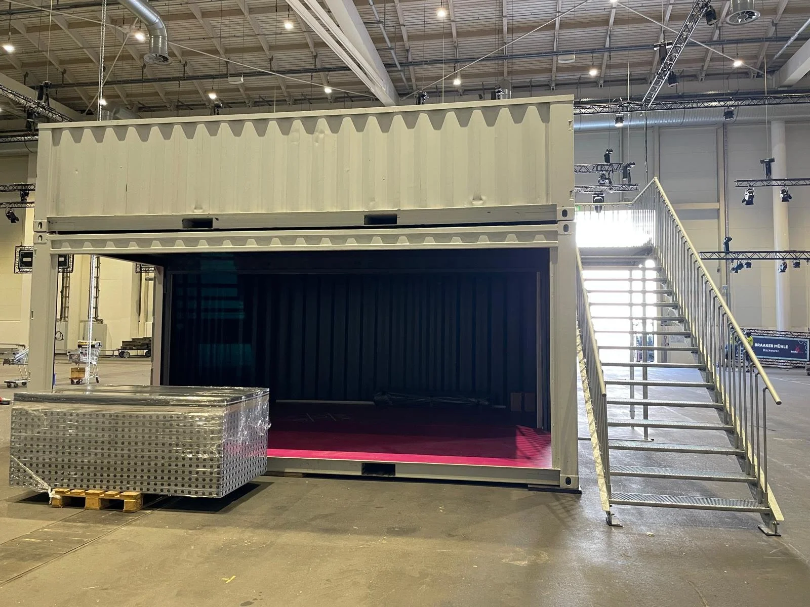 A large shipping container converted into a stage with stairs on the right side, inside a spacious industrial warehouse.