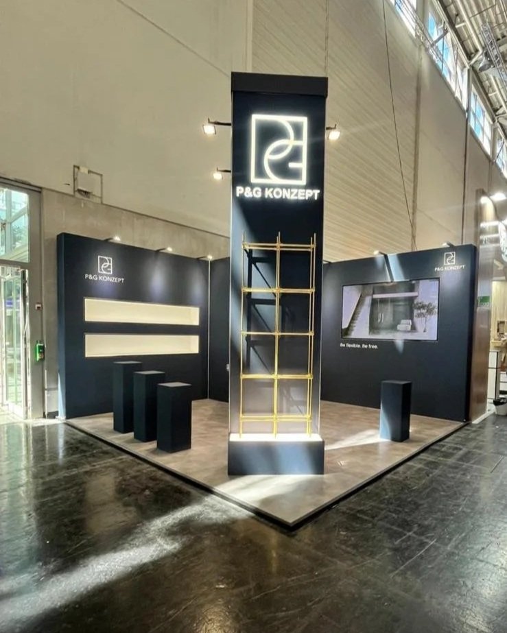 Trade show booth for P&G KONZEPT with black walls, a tall central display with a logo, seating area with three stools, and a TV screen on the right wall.