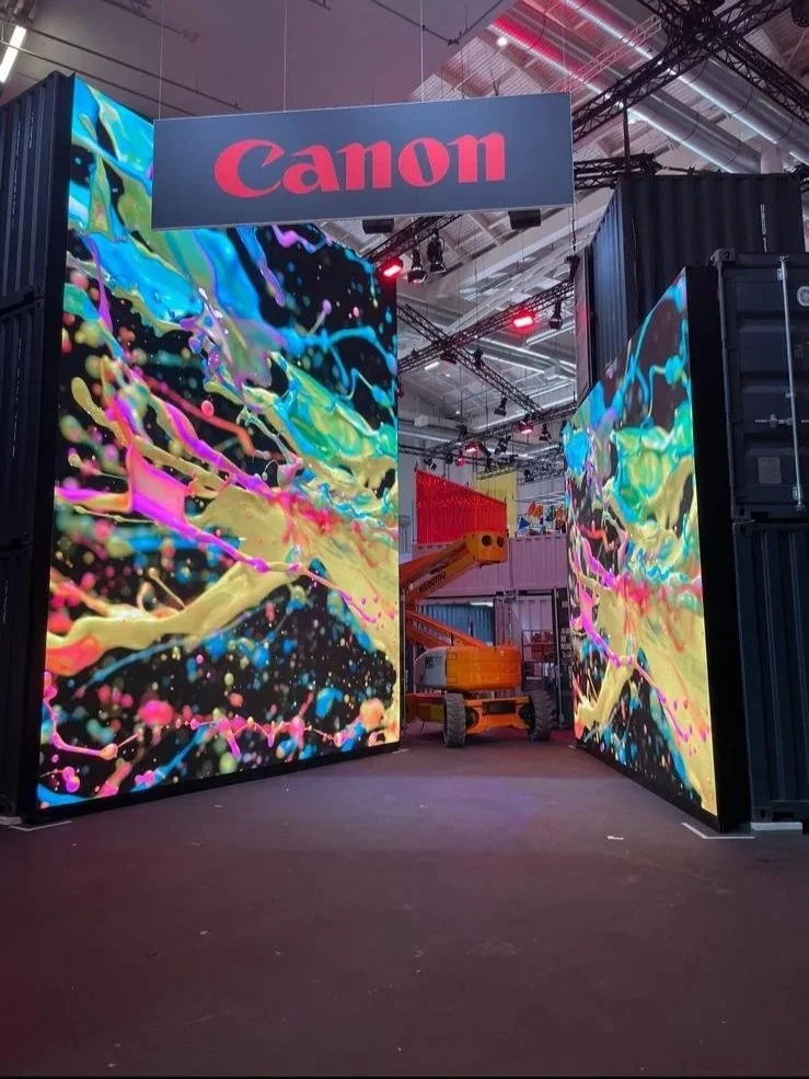 Large digital display screens at a Canon booth showcase colorful abstract artwork, with the Canon logo overhead at a technology or trade show.
