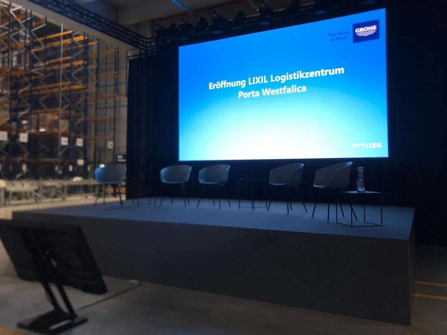 An empty stage in a warehouse with a large screen displaying text in German, a few chairs, and a water bottle on a table.