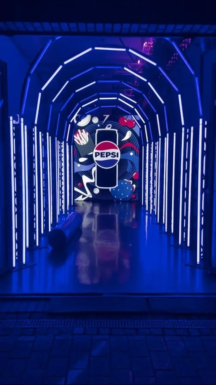 Neon-lit tunnel with Pepsi branding and graphics of cherries, fruit, and fries on the wall at the end of the hallway.