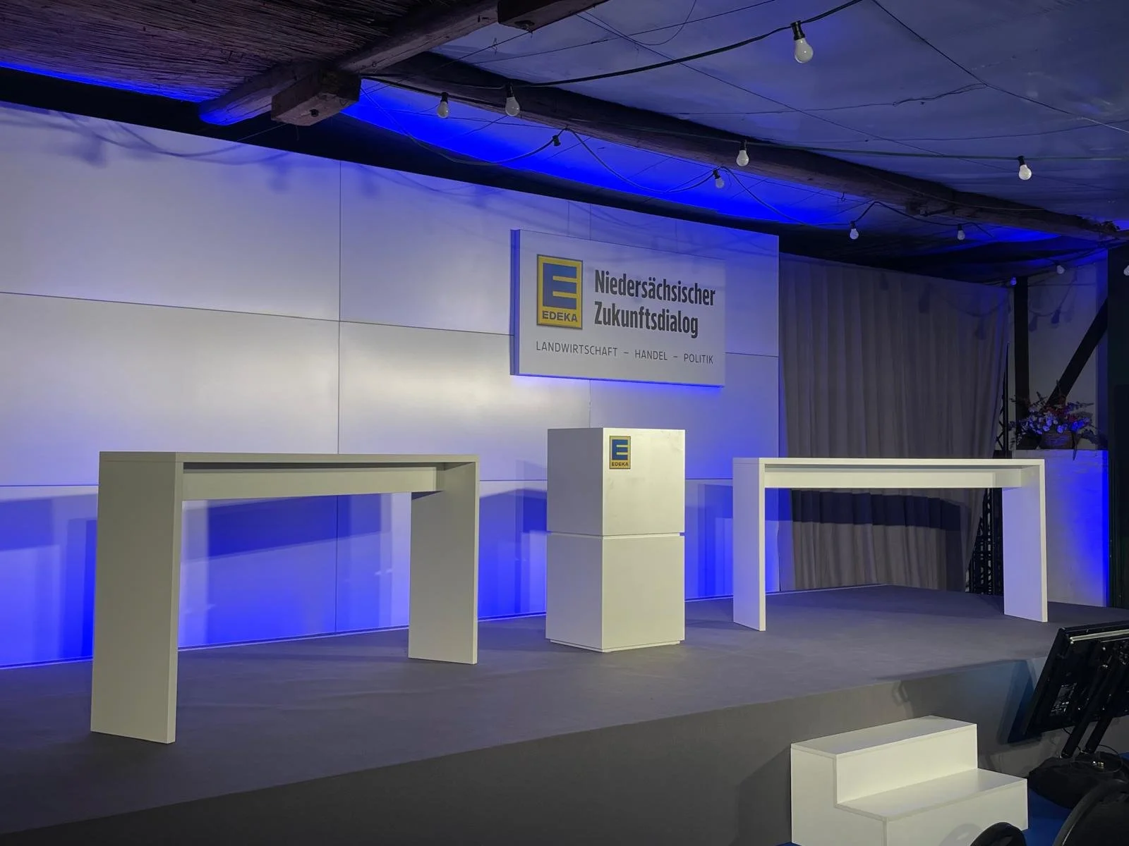 An empty stage set up with two white tables, a white podium with the EDEKA logo, and a large sign behind it reading 'Niedersächsischer Zukunftsdialog' with the EDEKA logo and the words 'Landwirtschaft - Handel - Politik,' illuminated by overhead ligh