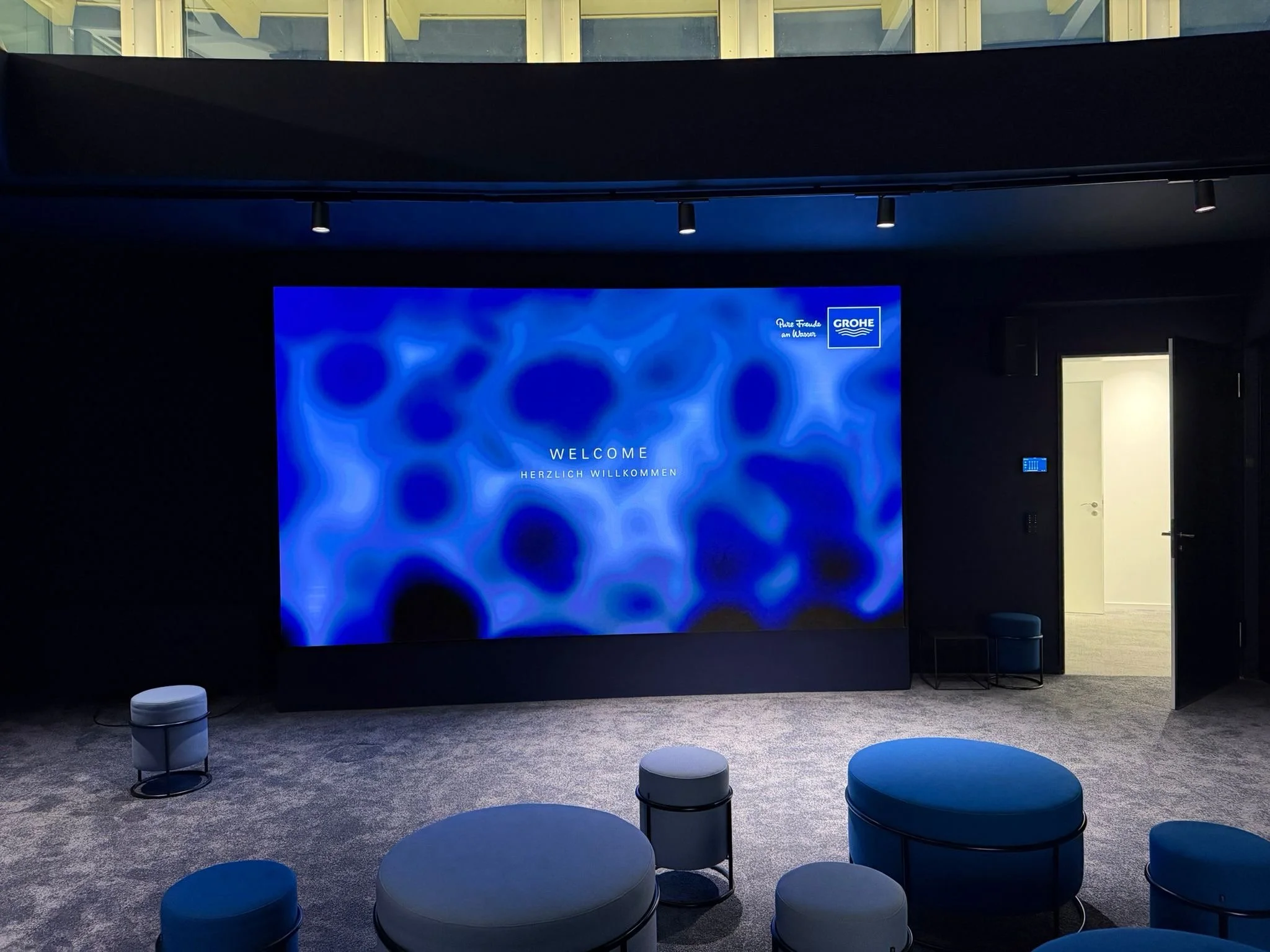A modern room with a large blue digital screen displaying a welcome message in English and German, with seating areas in front, and a door open to an adjacent white-walled space.