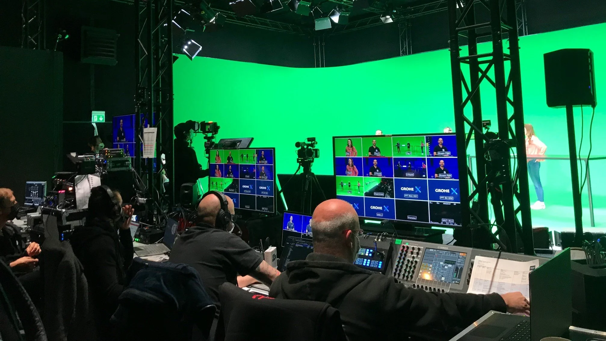 Behind the scenes of a green screen studio with multiple monitors, cameras, and technicians preparing for a broadcast or filming session.