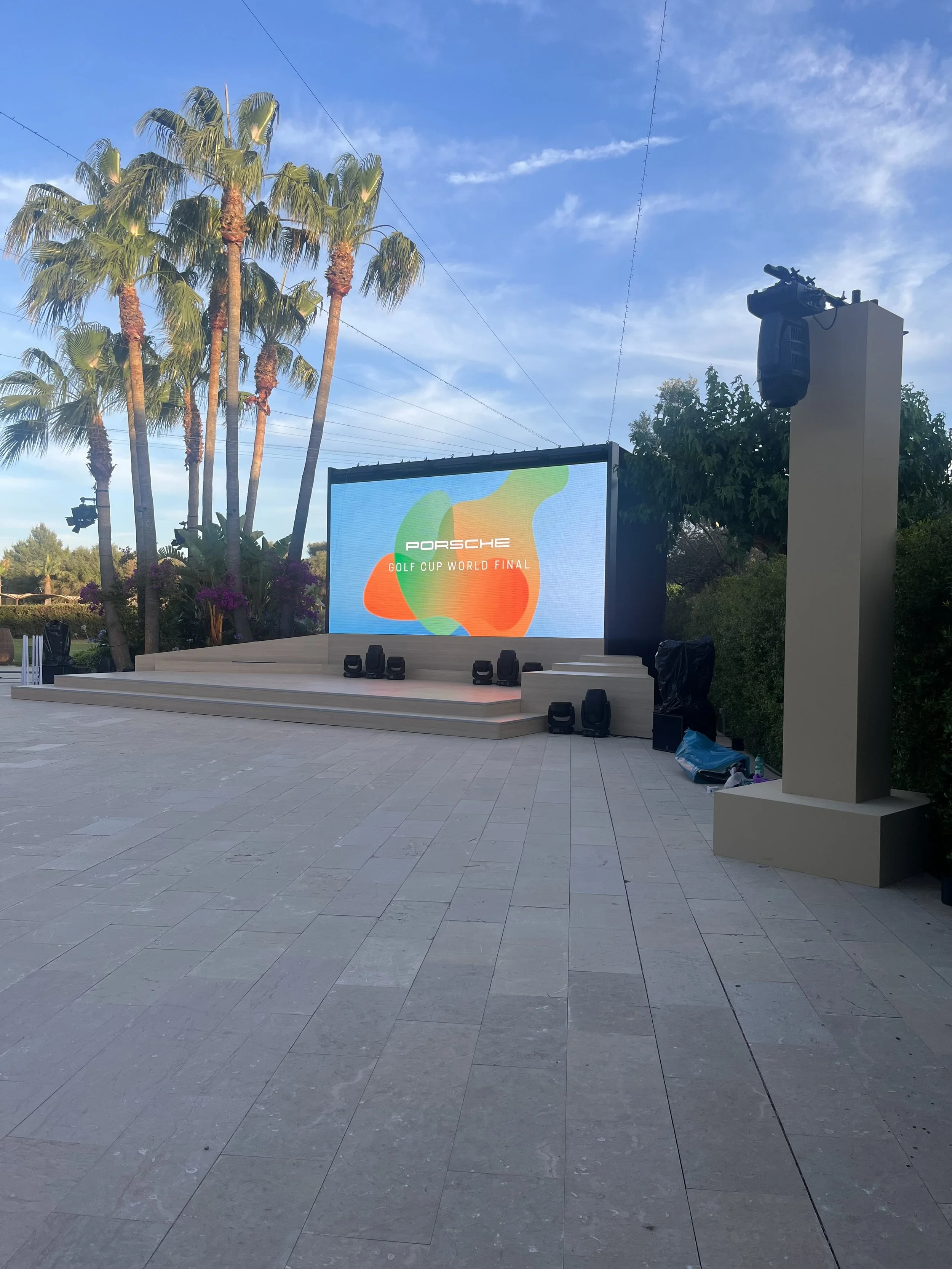 Outdoor stage with large screen displaying 'Porsche Golf Cup World Final' logo, surrounded by palm trees and set against a partly cloudy sky.