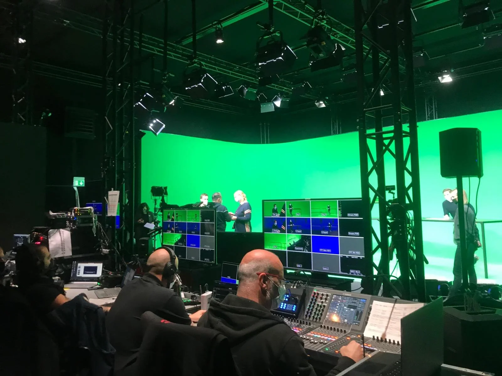 Broadcast control room with multiple screens and audio engineers working, behind a green screen being used for a live or recorded video production.