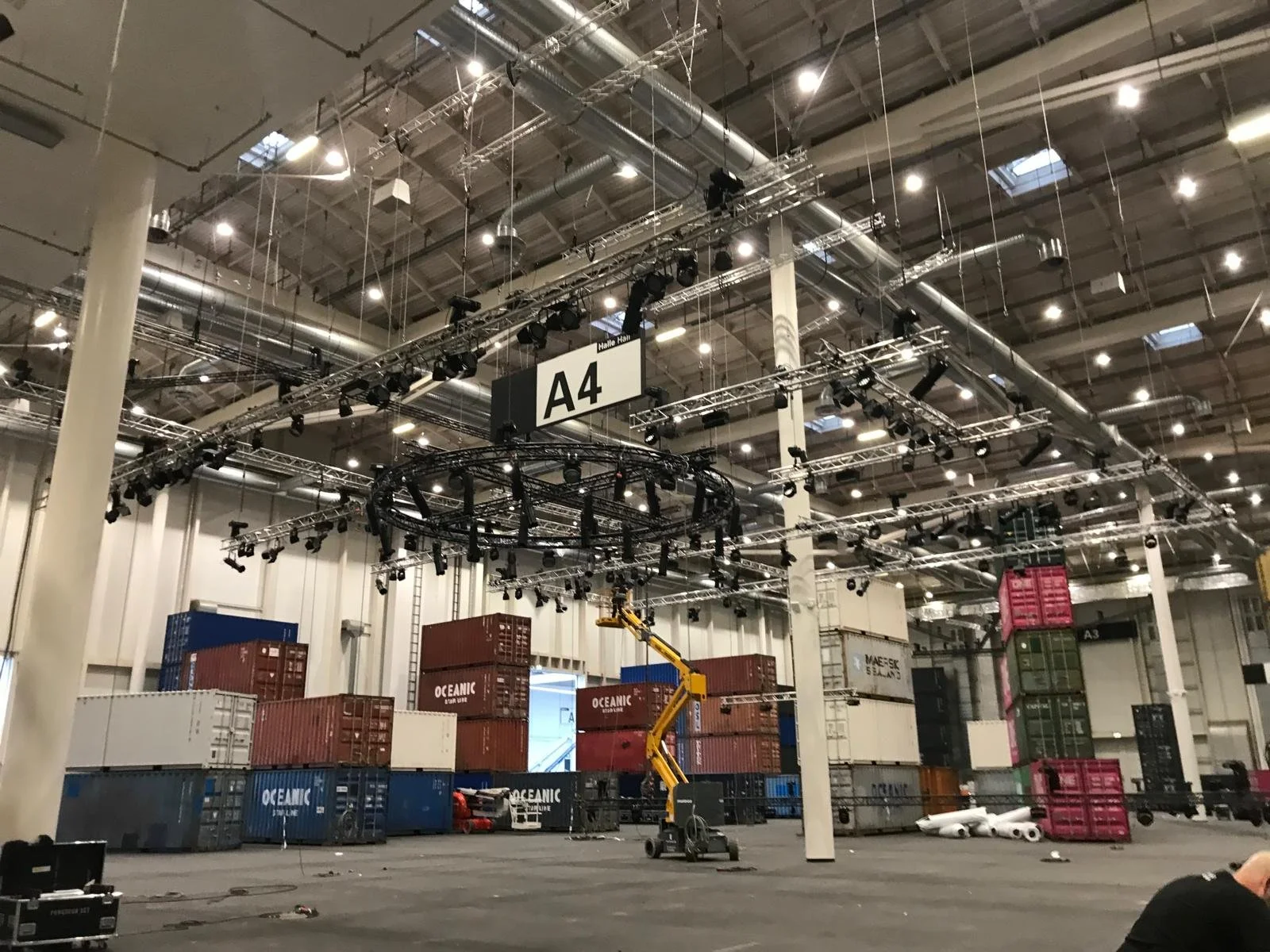 Interior of a large convention hall with numerous stacked shipping containers and a crane, illuminated by ceiling lights and equipped with a stage setup labeled A4.