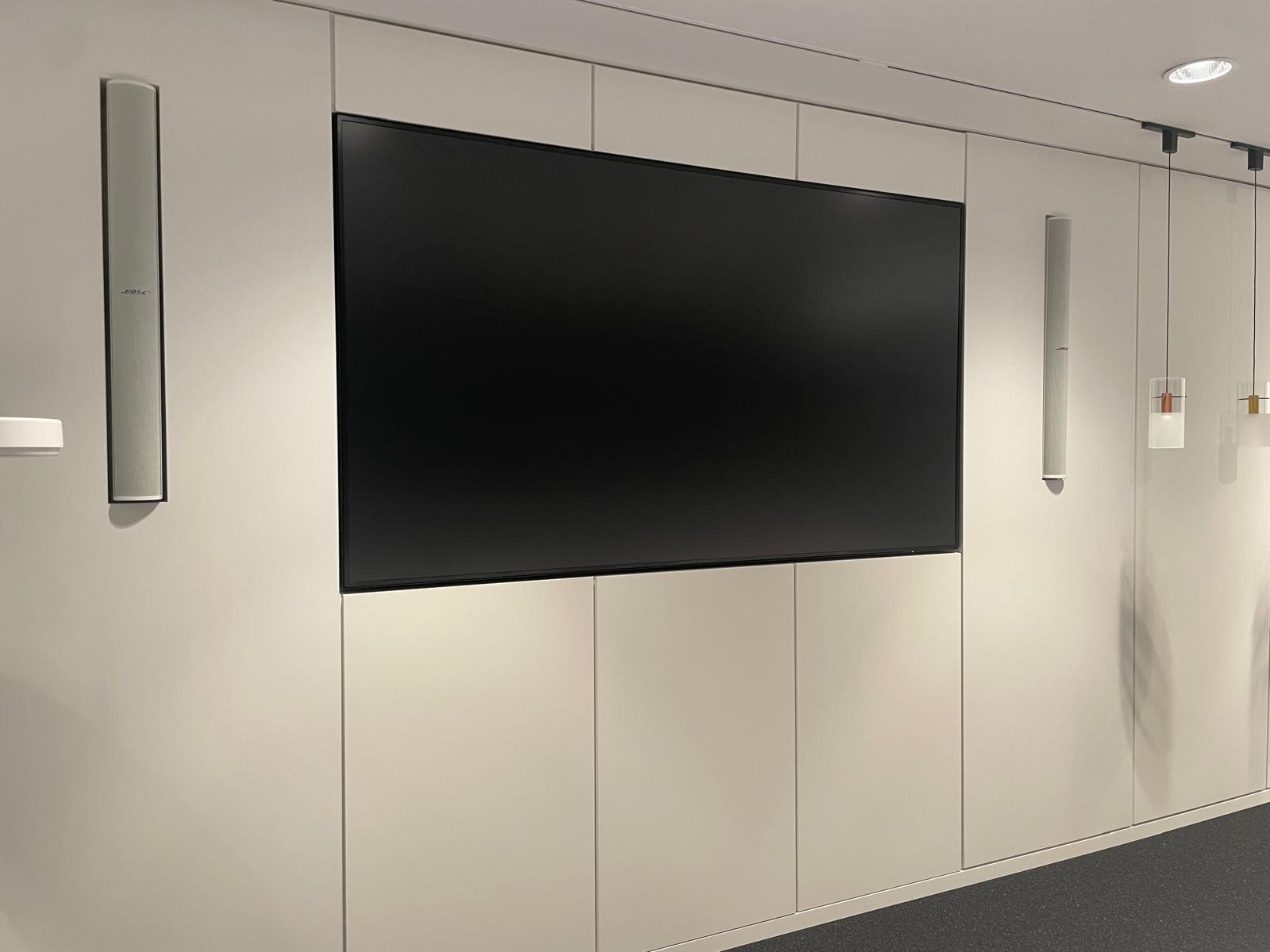 Modern conference room wall with a large flat-screen TV, two vertical speakers, and two small decorative wall lights.