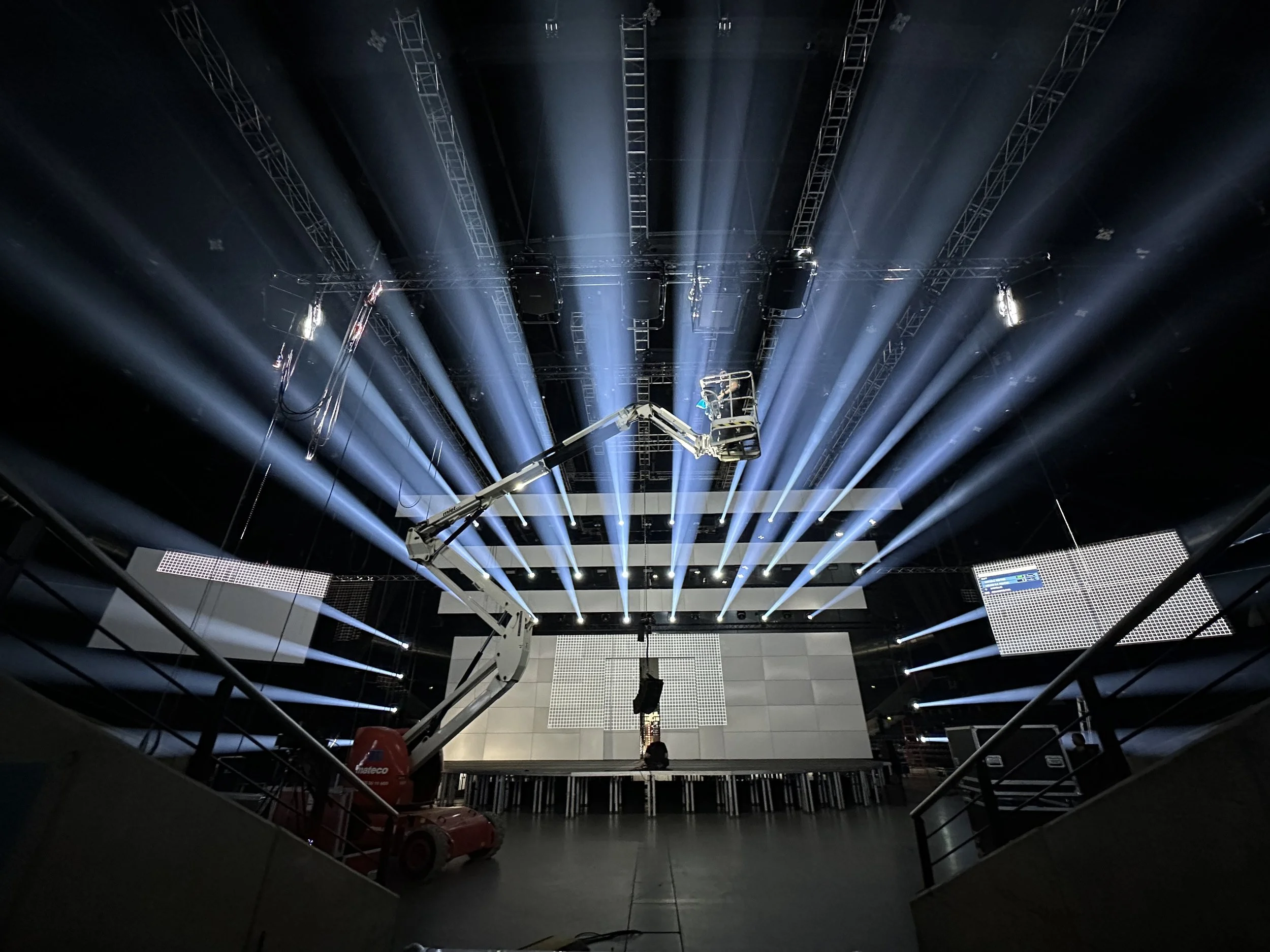 Inside a large event hall, a crane with a platform is positioned in the center, illuminated by multiple stage lights creating streaks of light across the ceiling ahead of large screens on stage.