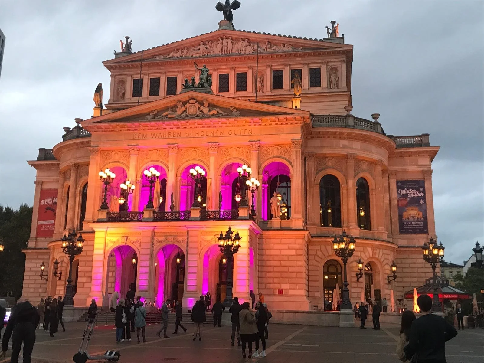 A grand, historic theater building illuminated with colorful pink and purple lights. The facade features ornate architectural details, statues, and balustrades. People are gathered outside on the plaza at dusk.