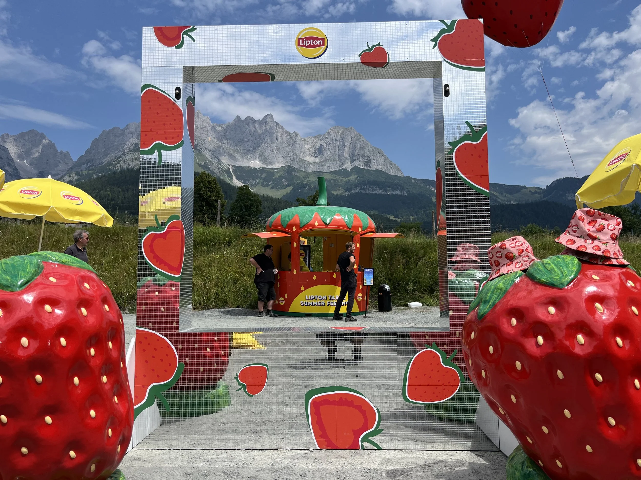 Outdoor scene with a mirrored photo frame decorated with strawberries and a portable strawberry-themed kiosk, set against a mountain backdrop with blue sky and clouds, and yellow umbrellas.
