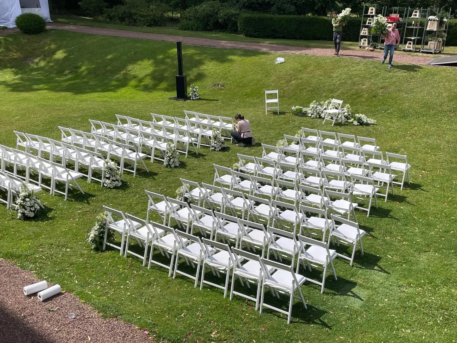 Outdoor wedding setup with white chairs arranged on green grass, floral arrangements at the ends and in the middle, and a person kneeling on the grass near the chairs. Two people are setting up flowers in the background, and there are shelves with de