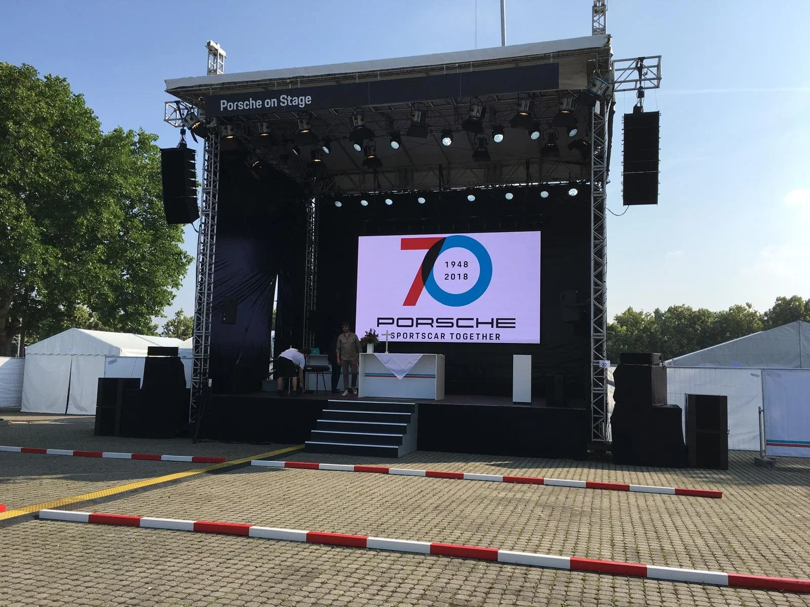 Outdoor stage with a large screen displaying the Porsche logo and a 70th anniversary celebration, surrounded by tents and trees.