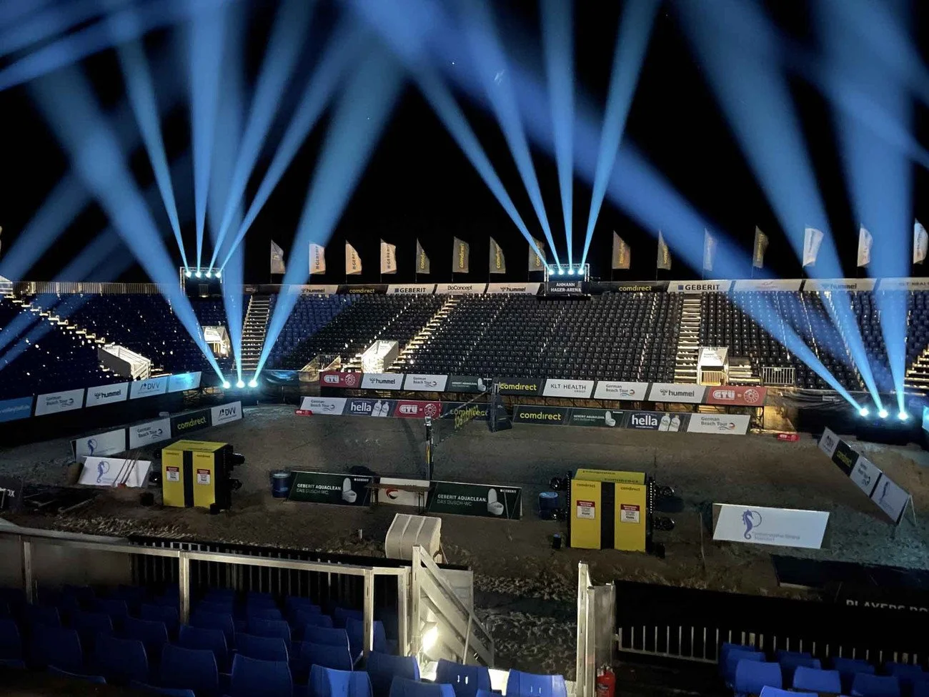 Empty outdoor stadium or arena lit with blue spotlights at night, surrounded by empty seats and advertising banners.