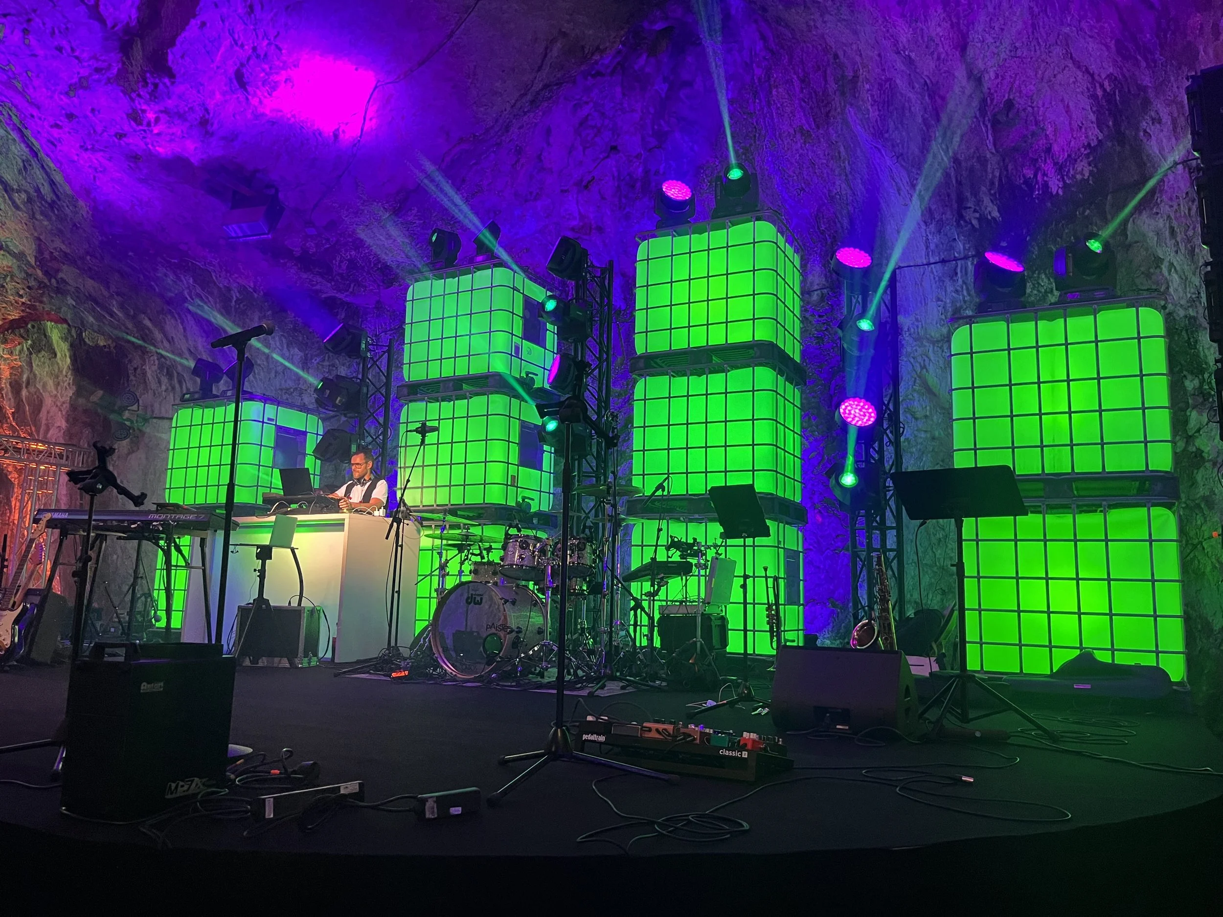 A stage set with bright green illuminated block structures, stage lights, musical instruments, and a DJ or musician in a cave-like environment.