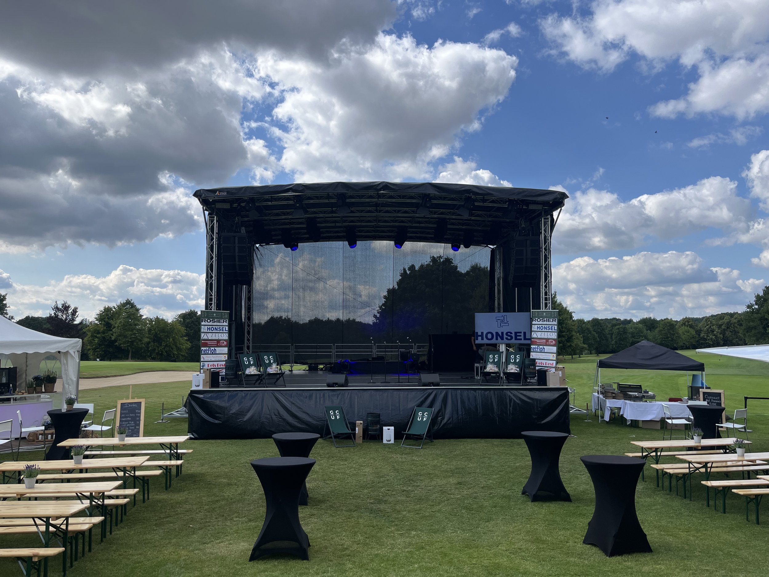 An outdoor stage set up on a grassy field with chairs and tables, under partly cloudy skies, likely for a concert or event.