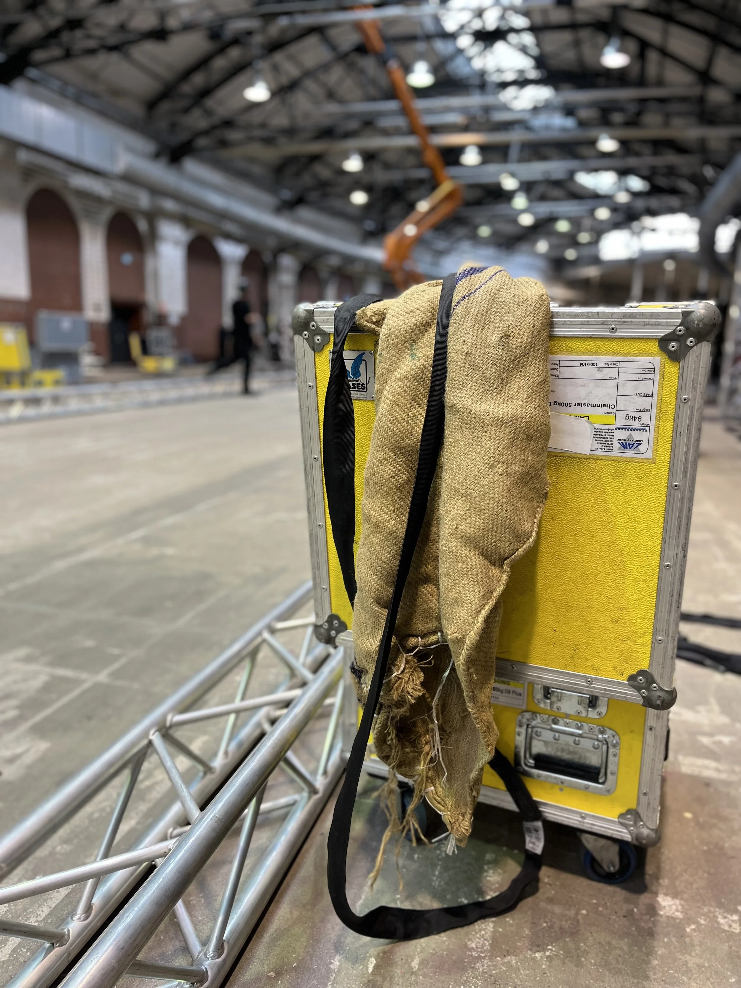 A yellow equipment case with a black strap and dirty rags hanging on it, located inside an industrial or warehouse space with high ceilings and visible structural beams.