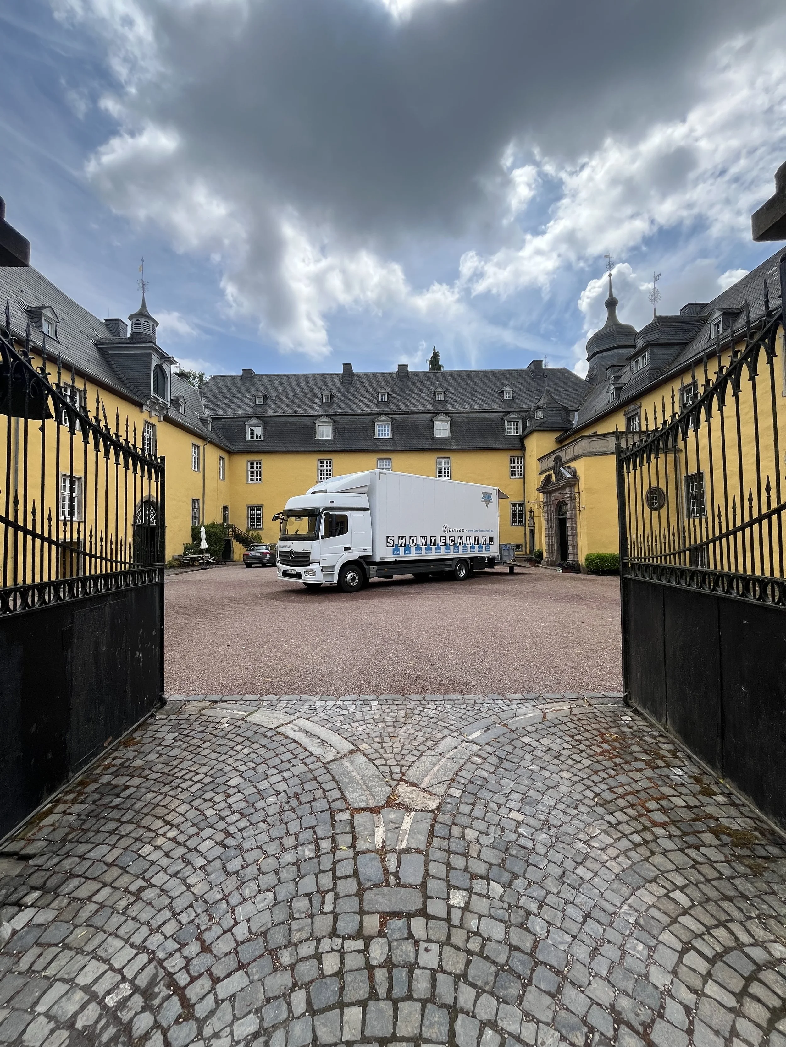 View through open gate of a courtyard with a large yellow building, a truck parked in the courtyard, and cloudy sky above.