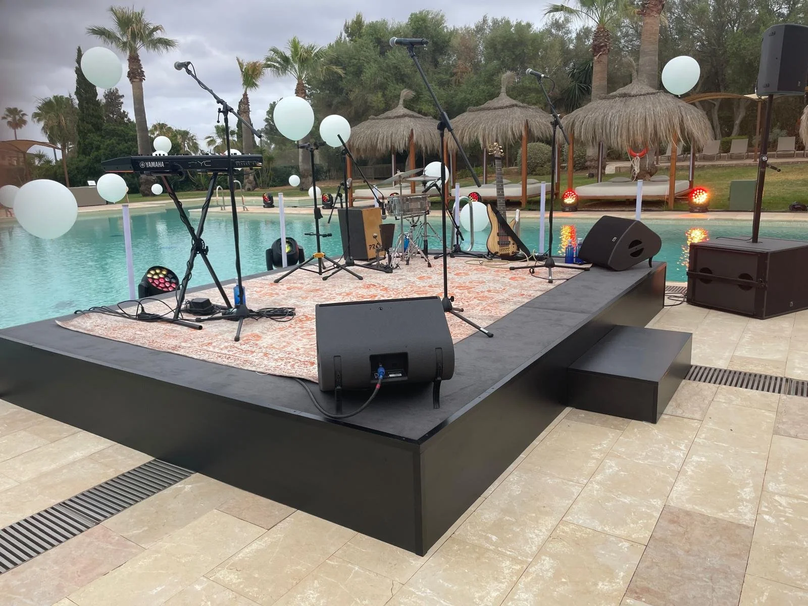 Music stage setup by a poolside with microphones, keyboard, guitar, and speakers before a cloudy sky.