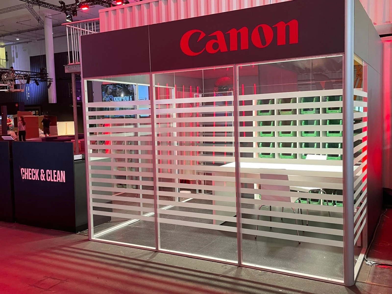 A Canon booth with a glass partition featuring horizontal white stripes, located in an indoor exhibition space. The booth has a sign that reads 'CHECK & CLEAN' on the front counter.