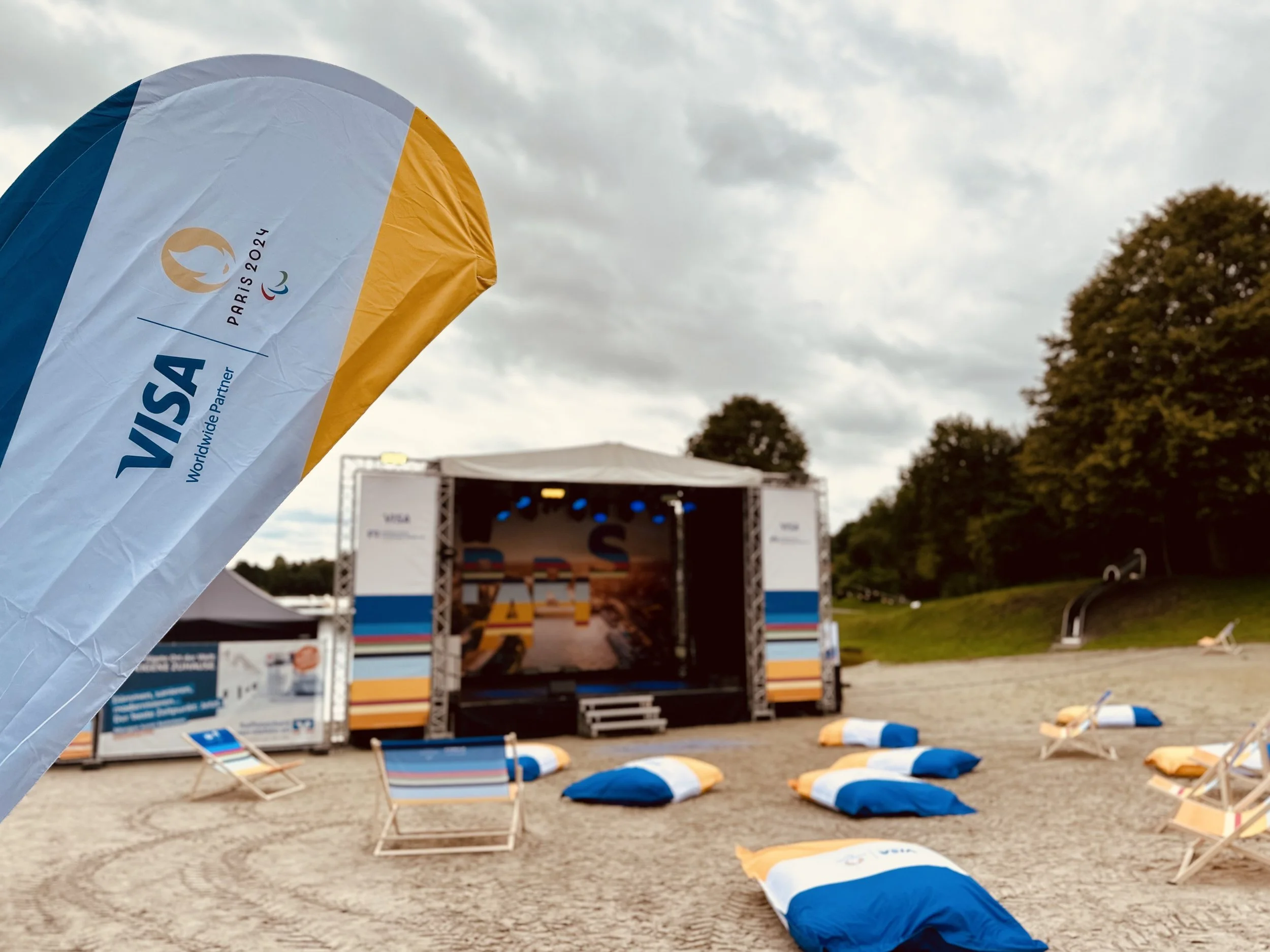 Outdoor stage setup with bean bag chairs and a large VIP flag promoting the Paris 2024 Olympics, with trees and cloudy sky in the background.