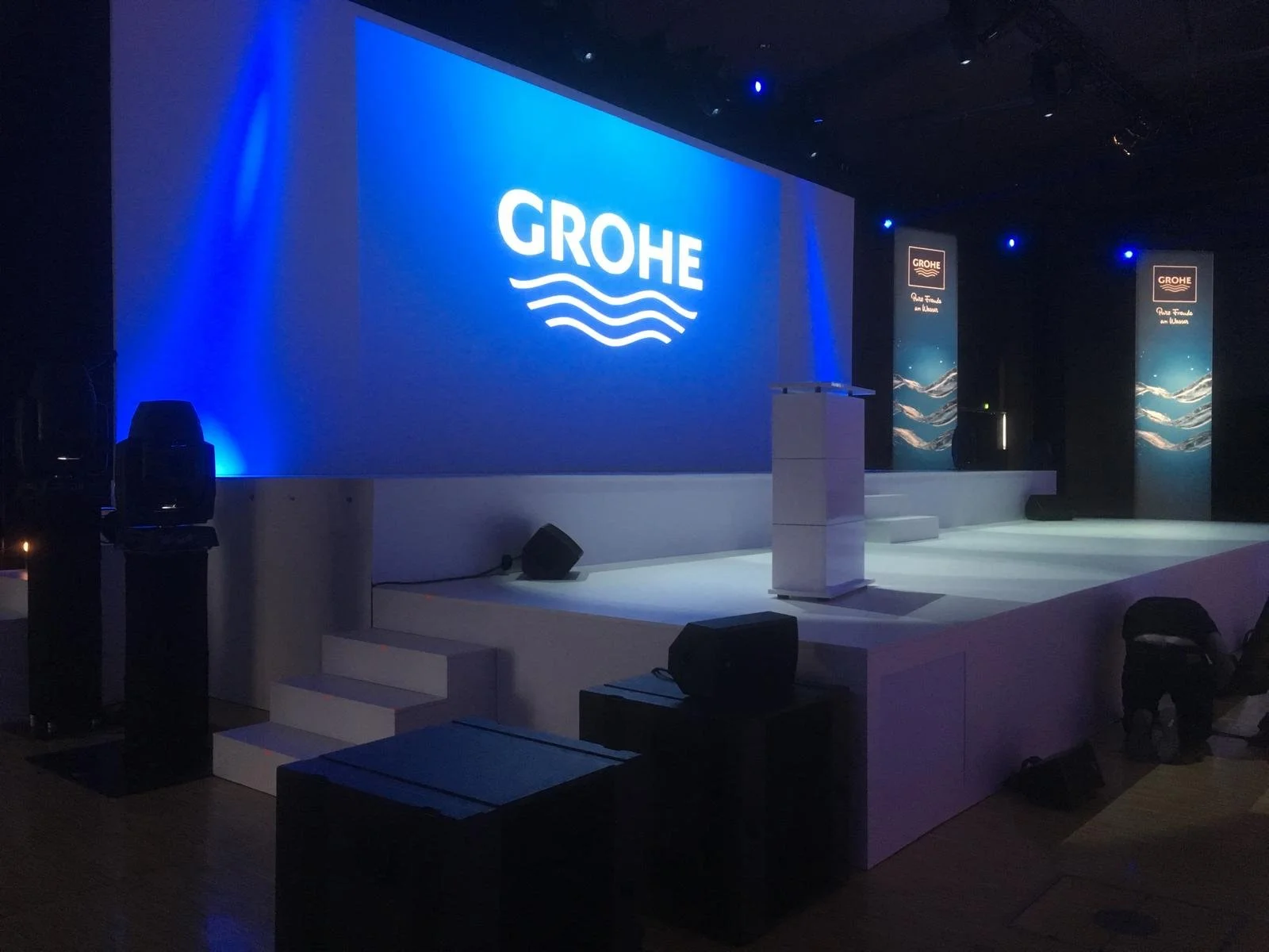 Stage set up with a large blue screen displaying the Grohe logo, surrounded by banners with the same branding, in a dark auditorium or conference hall.
