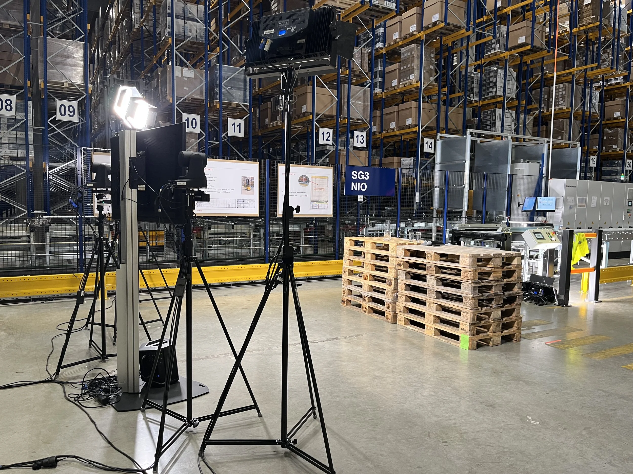 Interior of a warehouse with shelving units filled with boxes, a pallet with wooden pallets, and filming equipment including cameras, lights, and monitors set up for filming or photography.
