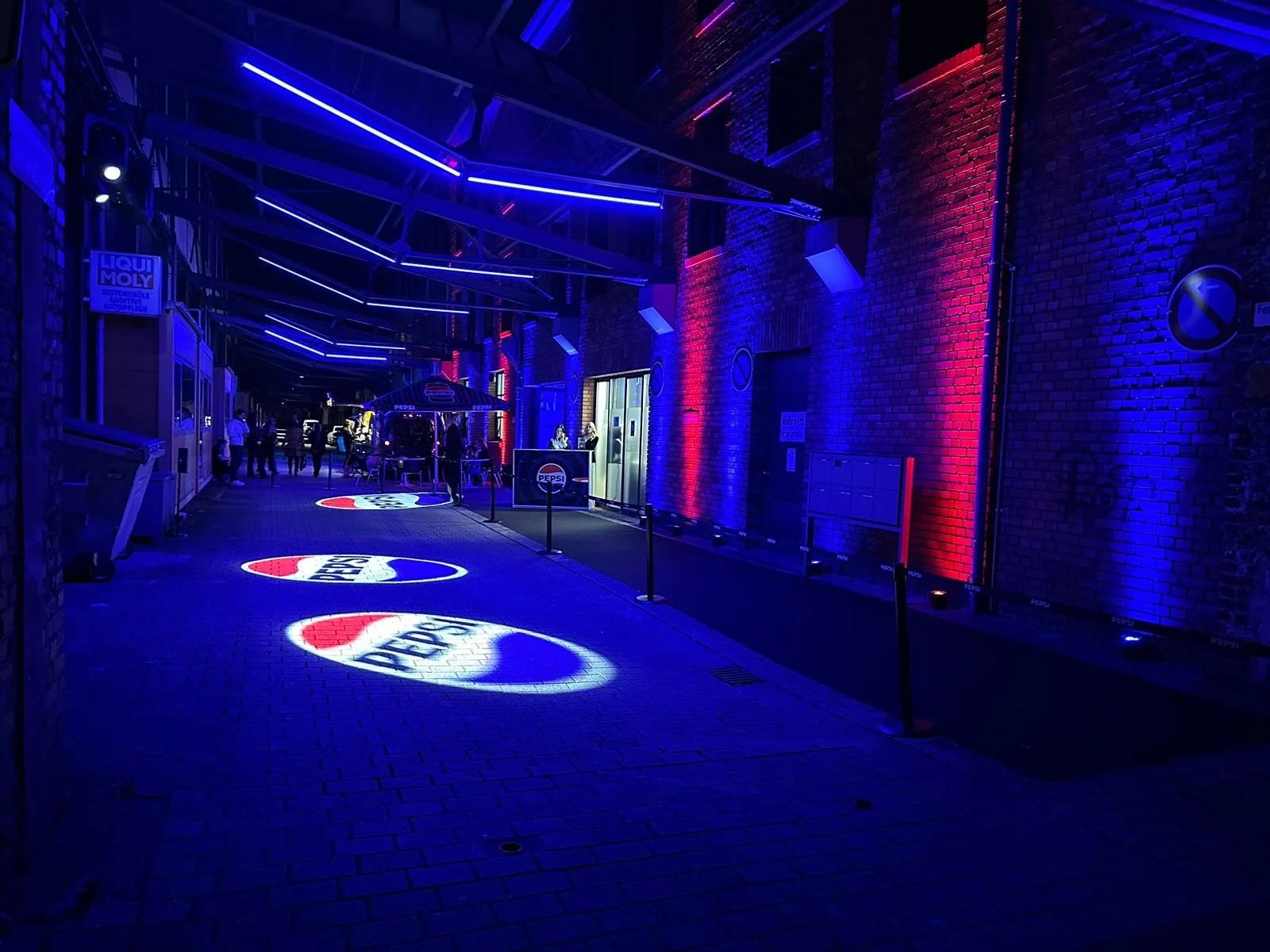 Nighttime scene of an outdoor bar with blue and red lighting, illuminated Pepsi logos on the ground, and people gathered around tables and the bar.