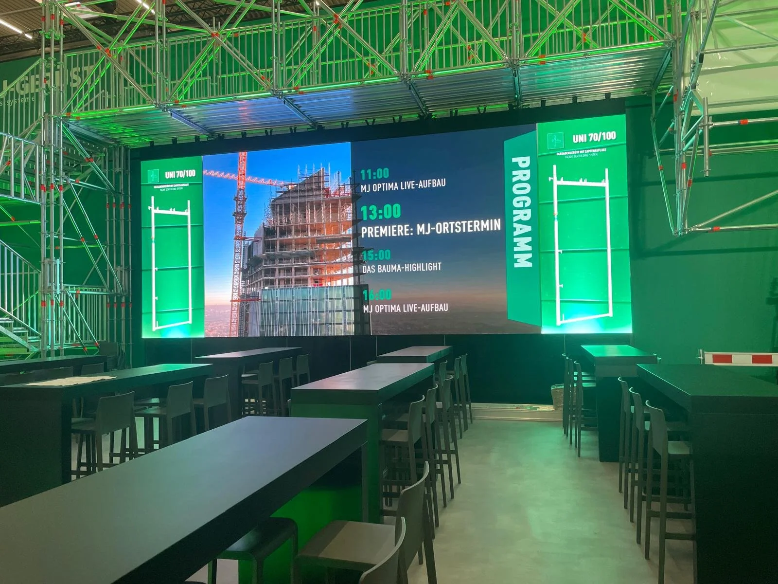 Indoor event space with a large digital timetable screen displaying an event schedule against a cityscape background. The space has green lighting, scaffoldings, and high tables with chairs.