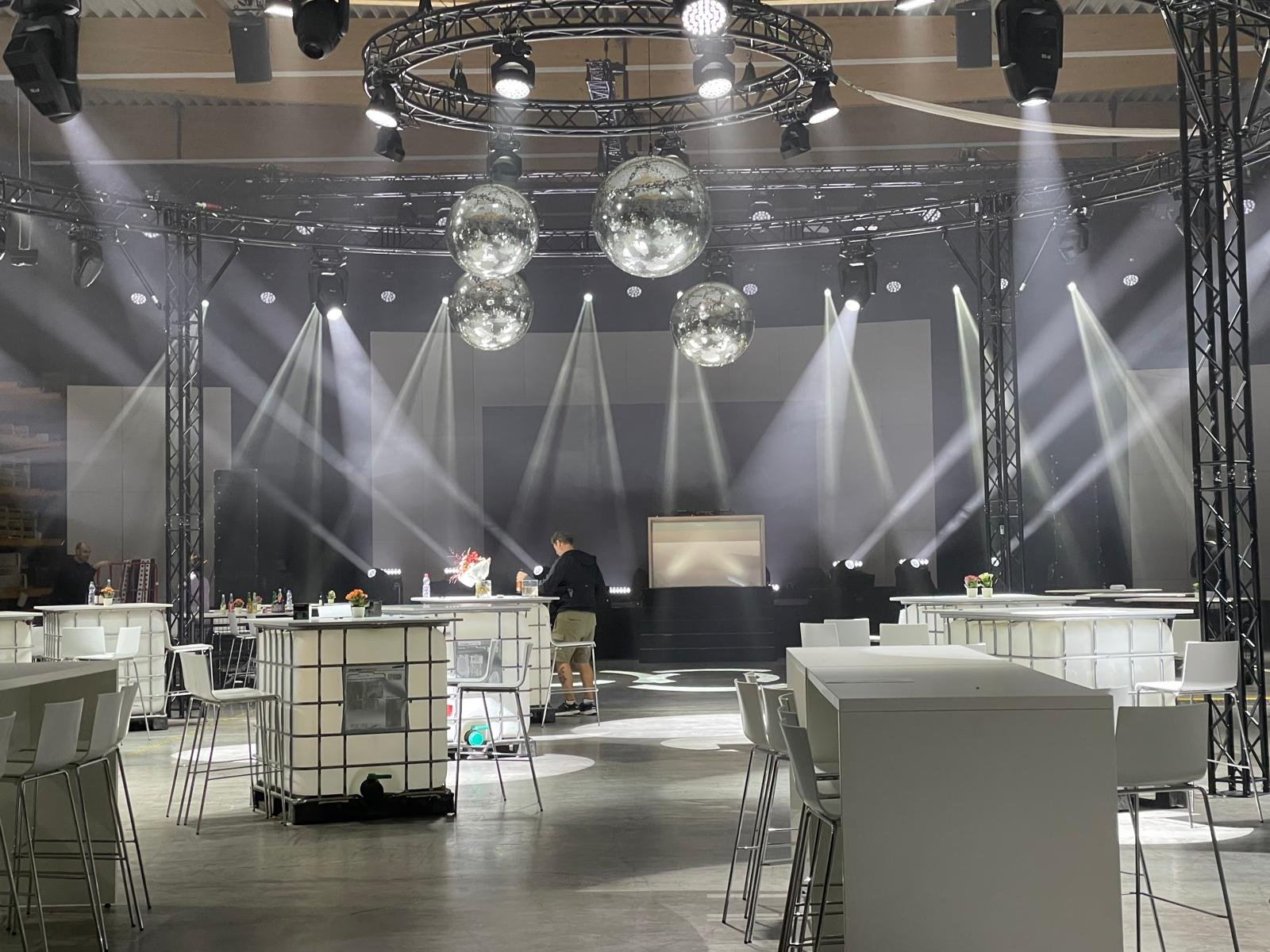 An indoor stage setup with bars and stools, bright stage lights, fog effects, and hanging decorative disco balls, suggesting preparations for a musical or entertainment event.