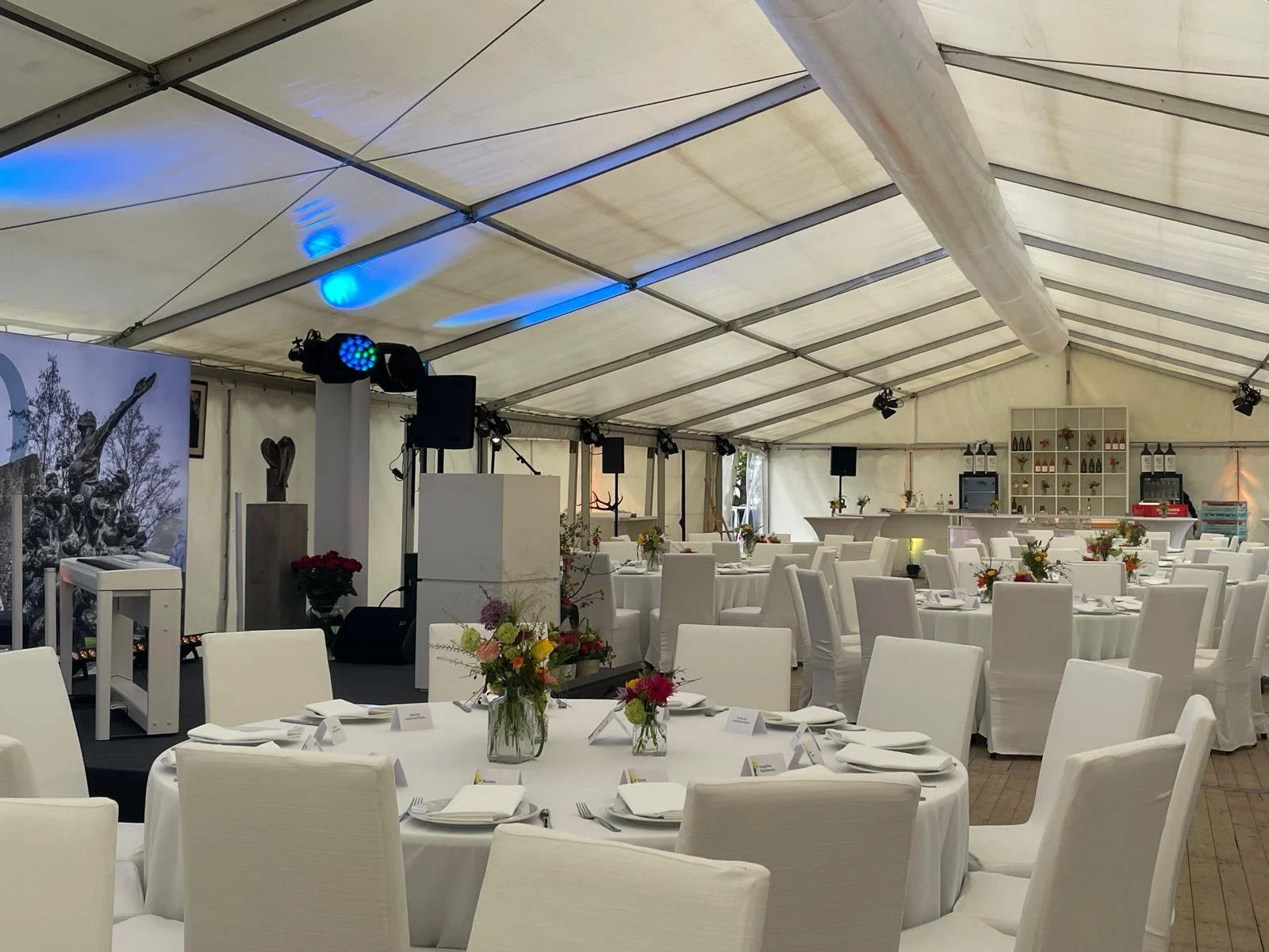 Inside a large event tent decorated for a formal gathering with round tables covered in white tablecloths, each set with plates, silverware, and folded napkins, and floral centerpieces. The background shows a bar area with shelves holding bottles and