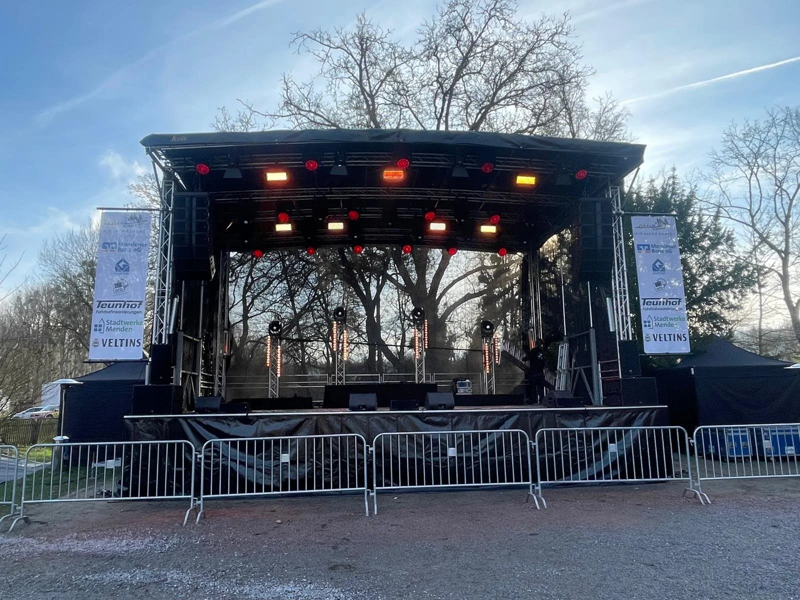 Empty outdoor stage with lighting and sound equipment, metal barriers in front, and banners on sides, set up for an event, with leafless trees and a partly cloudy sky in the background.