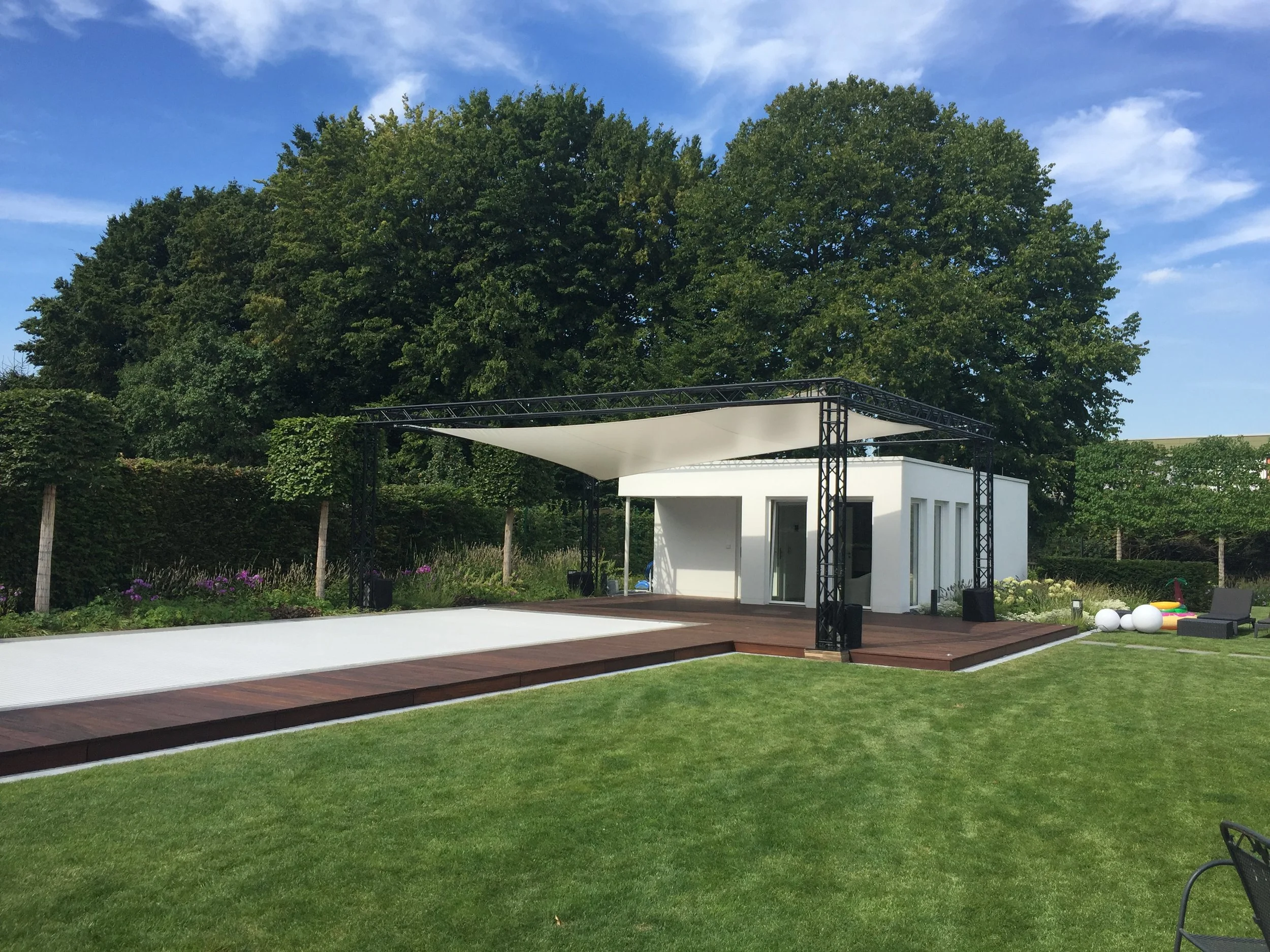 Modern backyard with a small white building, a wooden deck, a shade sail, lush green grass, and outdoor seating, surrounded by trees and a fence.