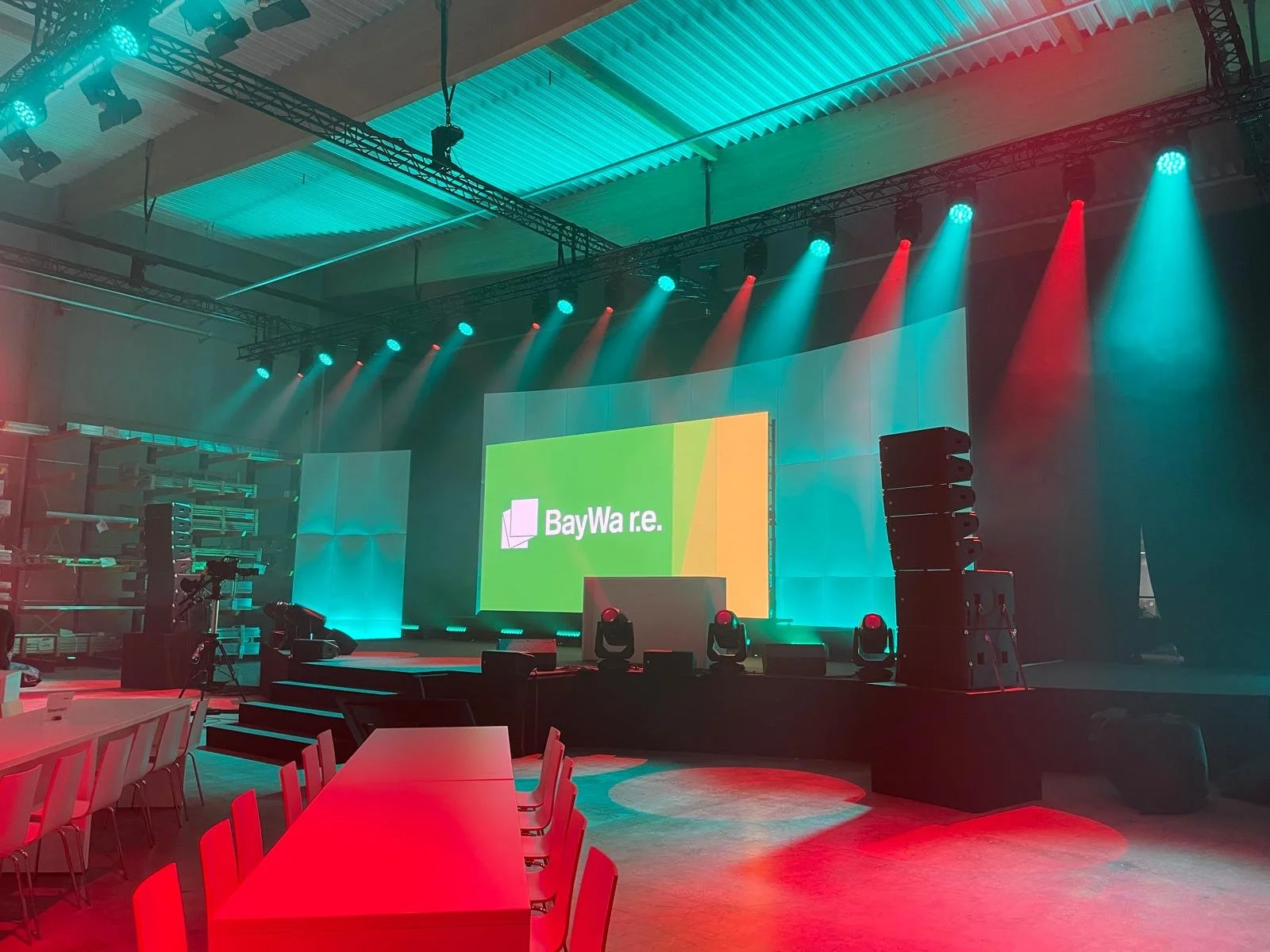Stage with large screen displaying 'BayWa re.' logo, surrounded by colored stage lights, in an indoor event space with seating and tables.