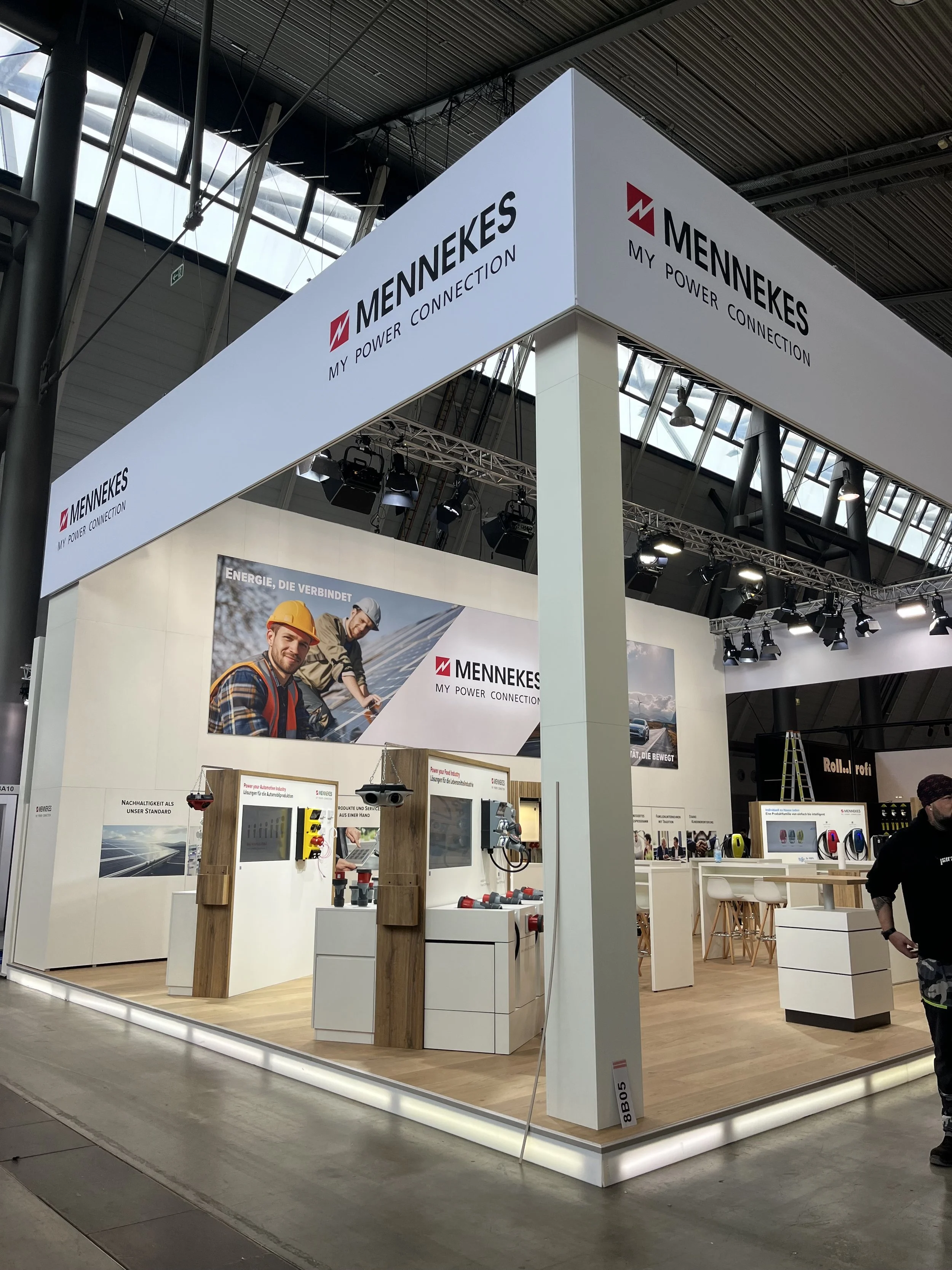 Trade show booth for MENNEKES with displays of electrical components, promotional posters, and people attending at an indoor exhibition hall.