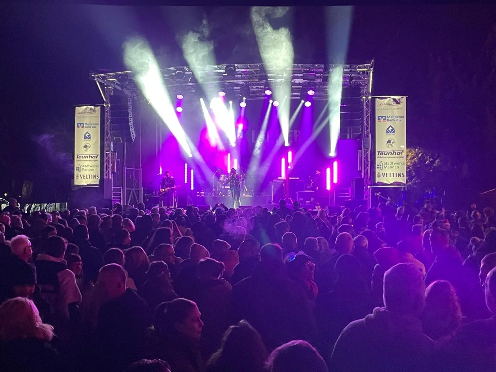 Nighttime outdoor concert with a large crowd watching a band perform on stage illuminated by purple and white lights.