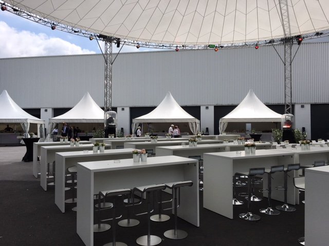 Outdoor event setup with white high tables and bar stools under a large canopy and tents, with a few people preparing or setting up.