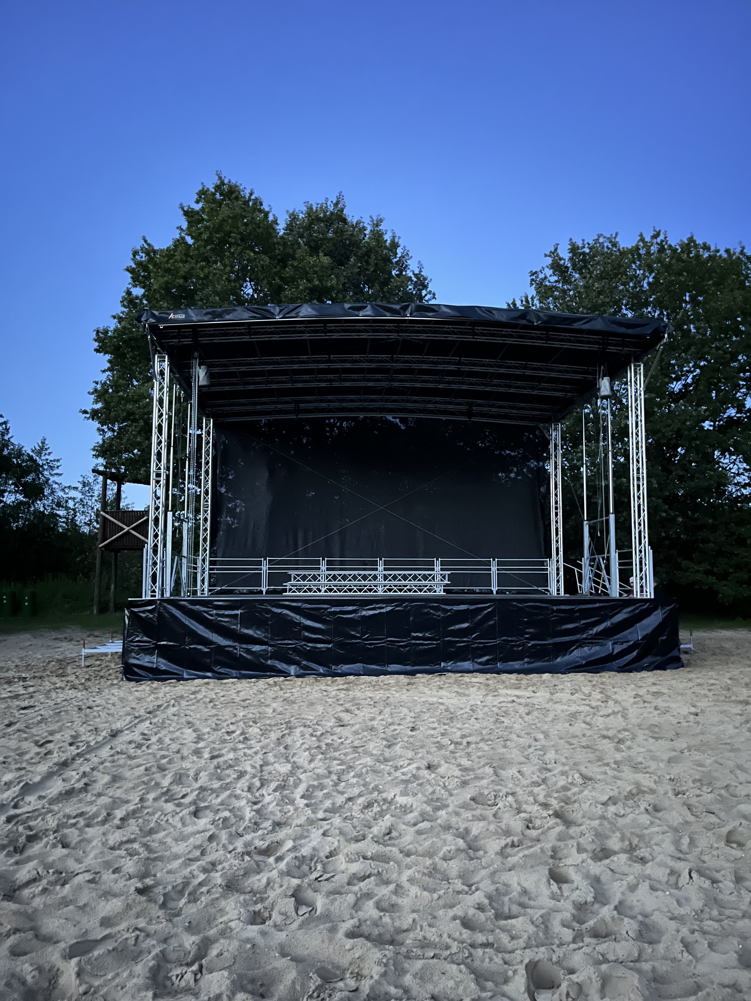 Empty outdoor stage with black backdrop and metal framework on sandy ground, surrounded by trees at dusk.