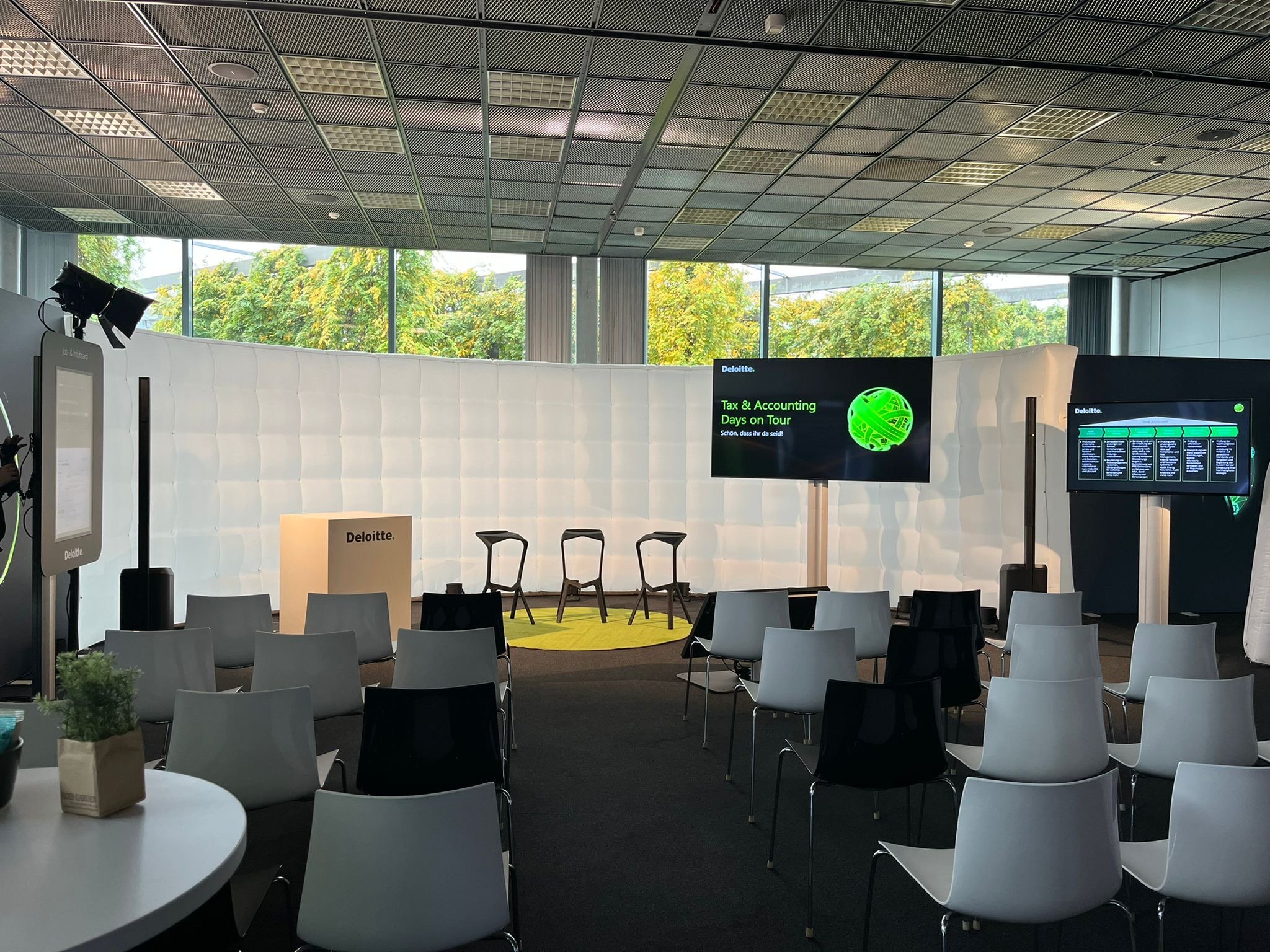 Conference room or event space with chairs arranged in rows, large screens displaying presentation titles, a white cushioned wall, and a window with trees outside.