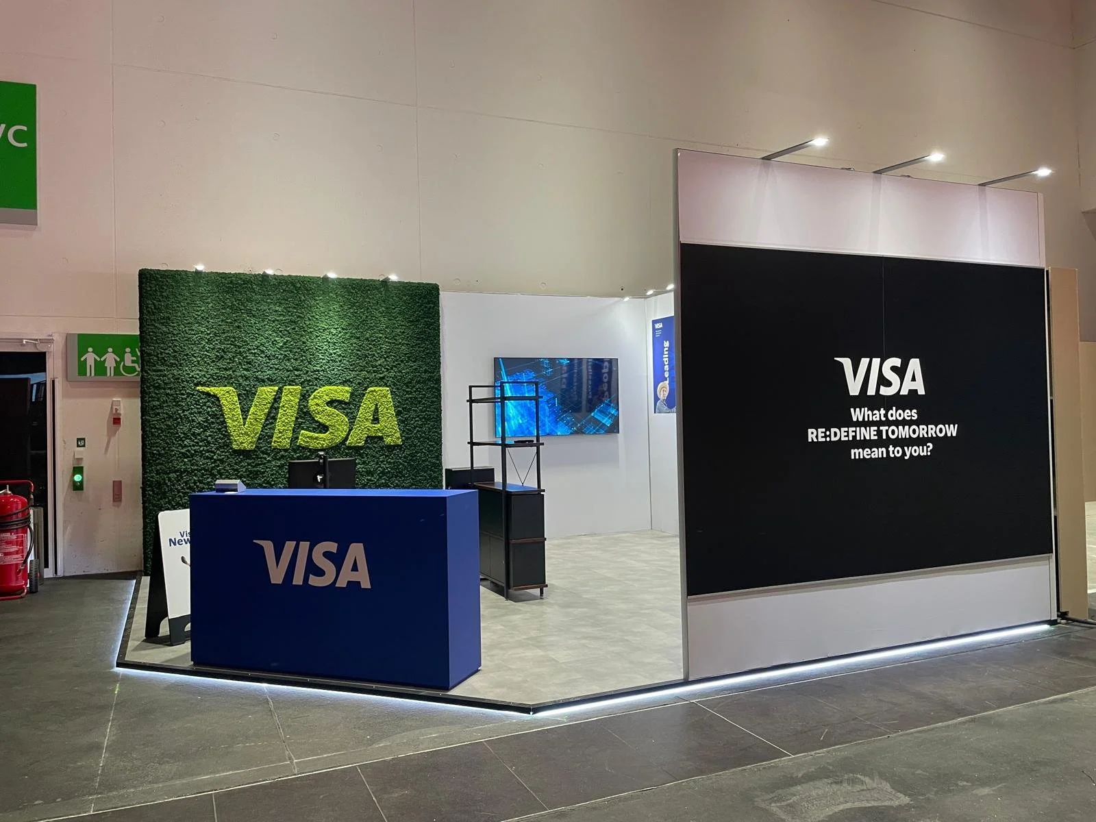 VISA booth with a green wall displaying the VISA logo, a blue counter with VISA branding, a large black sign with VISA logo and the question 'What does RE:DEFINE TOMORROW mean to you?', and a TV screen on a white wall.