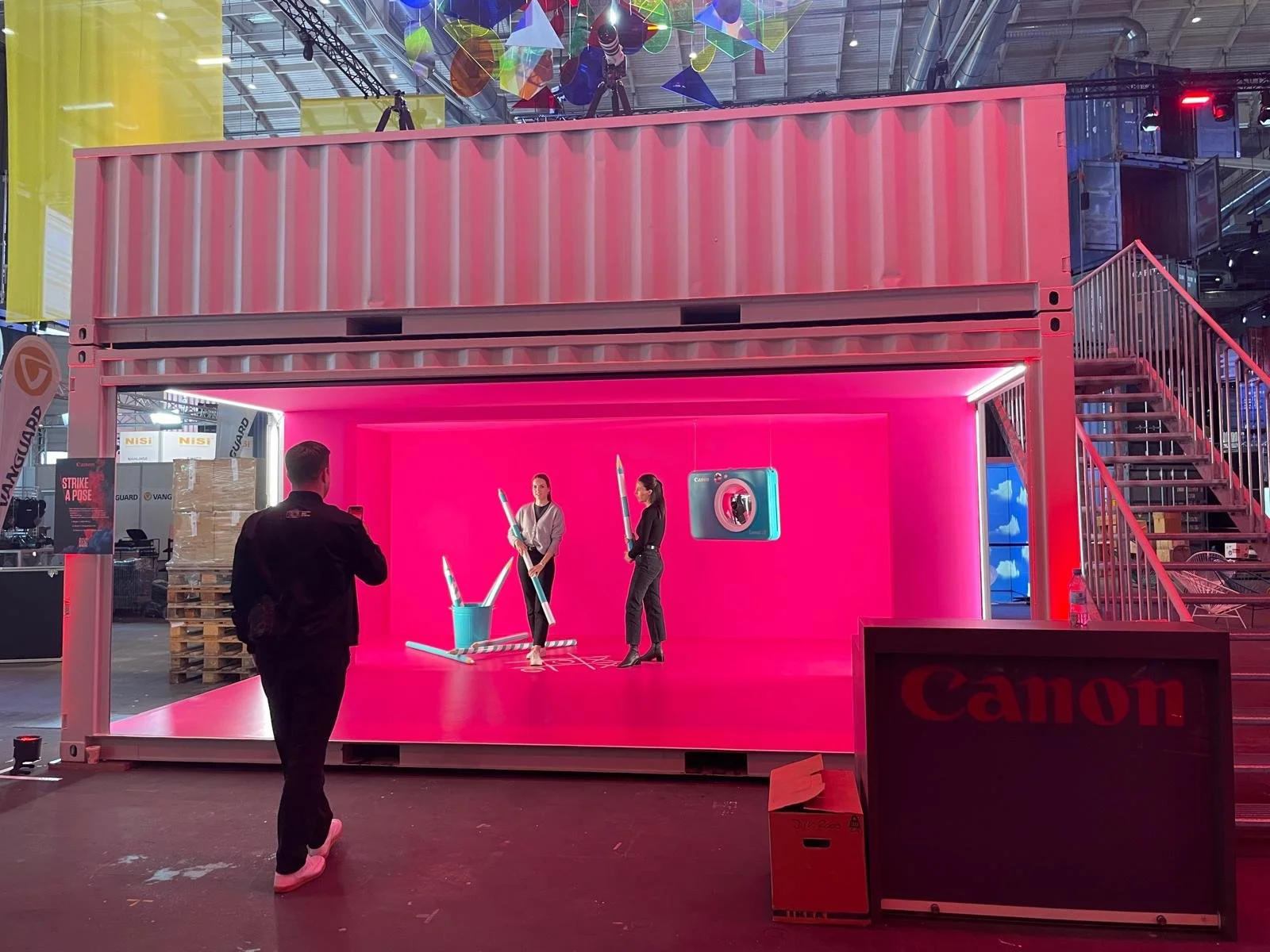 A trade show booth with a pink backdrop inside a container structure, displaying promotional images and a camera. Two women hold large pens, while a man in a black jacket with a Canon logo on the back stands in front, taking a photo. The booth is lab