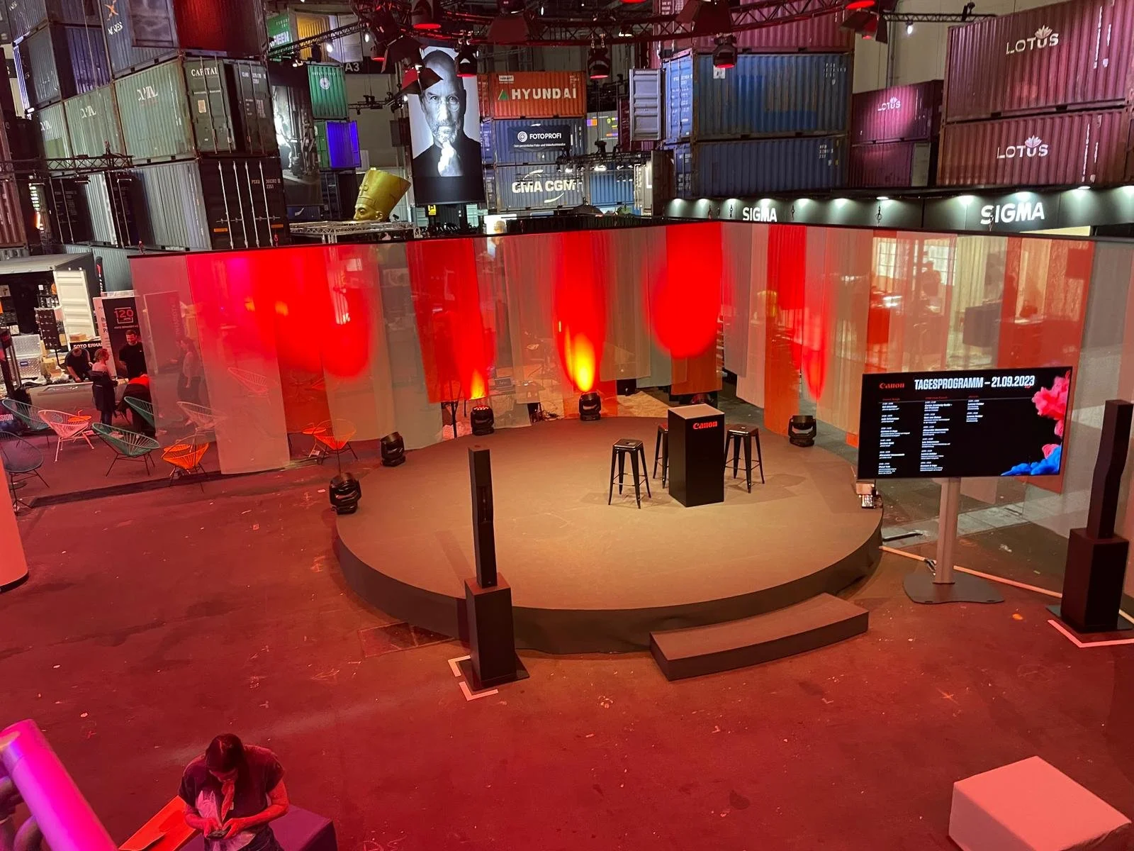 A round stage with three black stools, a Canon-branded podium, and a large digital screen displaying an event schedule, surrounded by orange and beige curtains with red and yellow lighting, inside a venue with stacked shipping containers and colorful